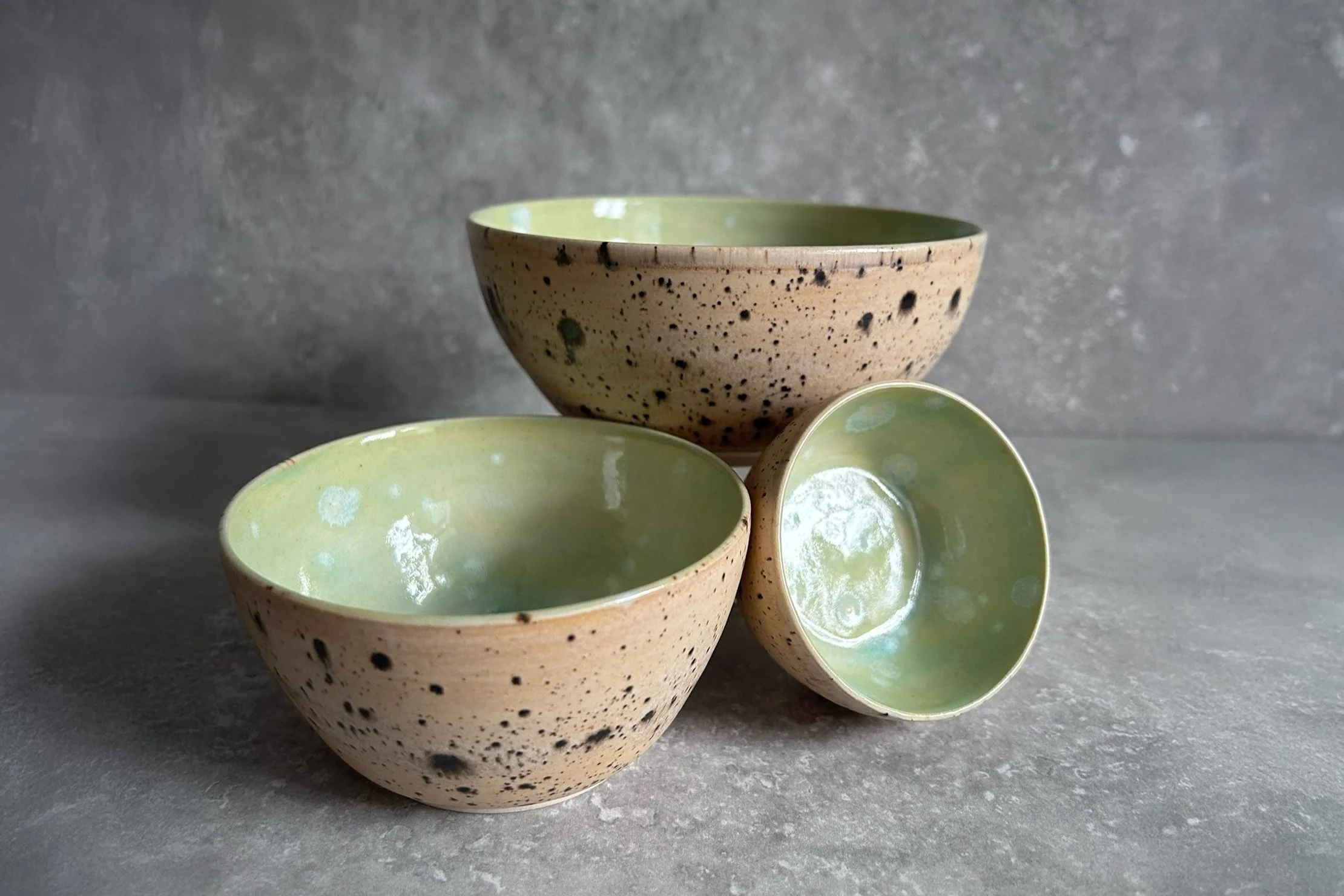 Three ceramic stacking bowls, two with a brown exterior speckled with black, and green glazed interiors, arranged on a textured gray surface against a gray background.