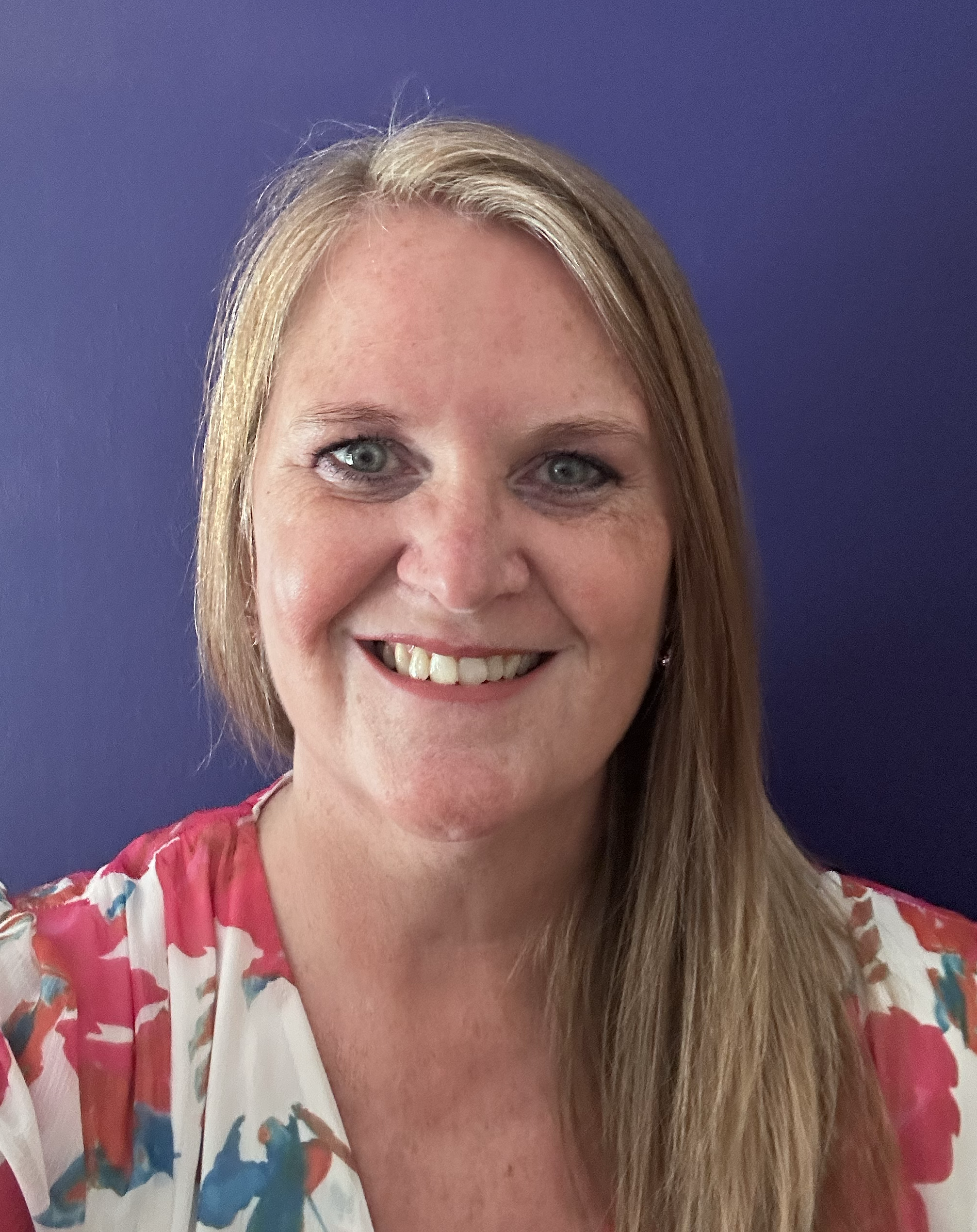 A smiling woman with strawberry blonde hair, wearing a floral blouse, standing in front of a plain purple wall.
