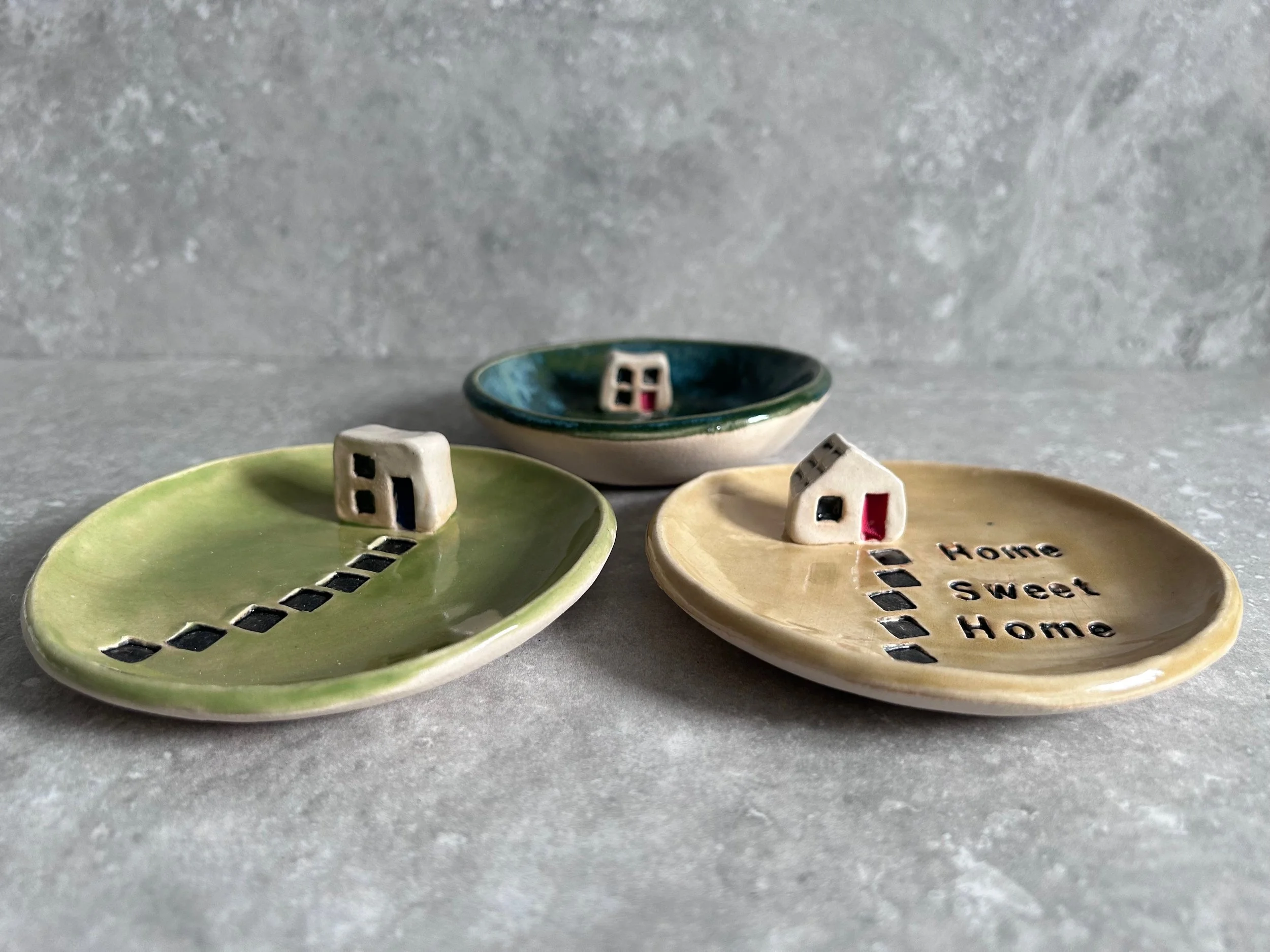 Three small ceramic dish plates with miniature house and pathway designs, one with the words 'Home Sweet Home' written on it, on a textured gray surface.