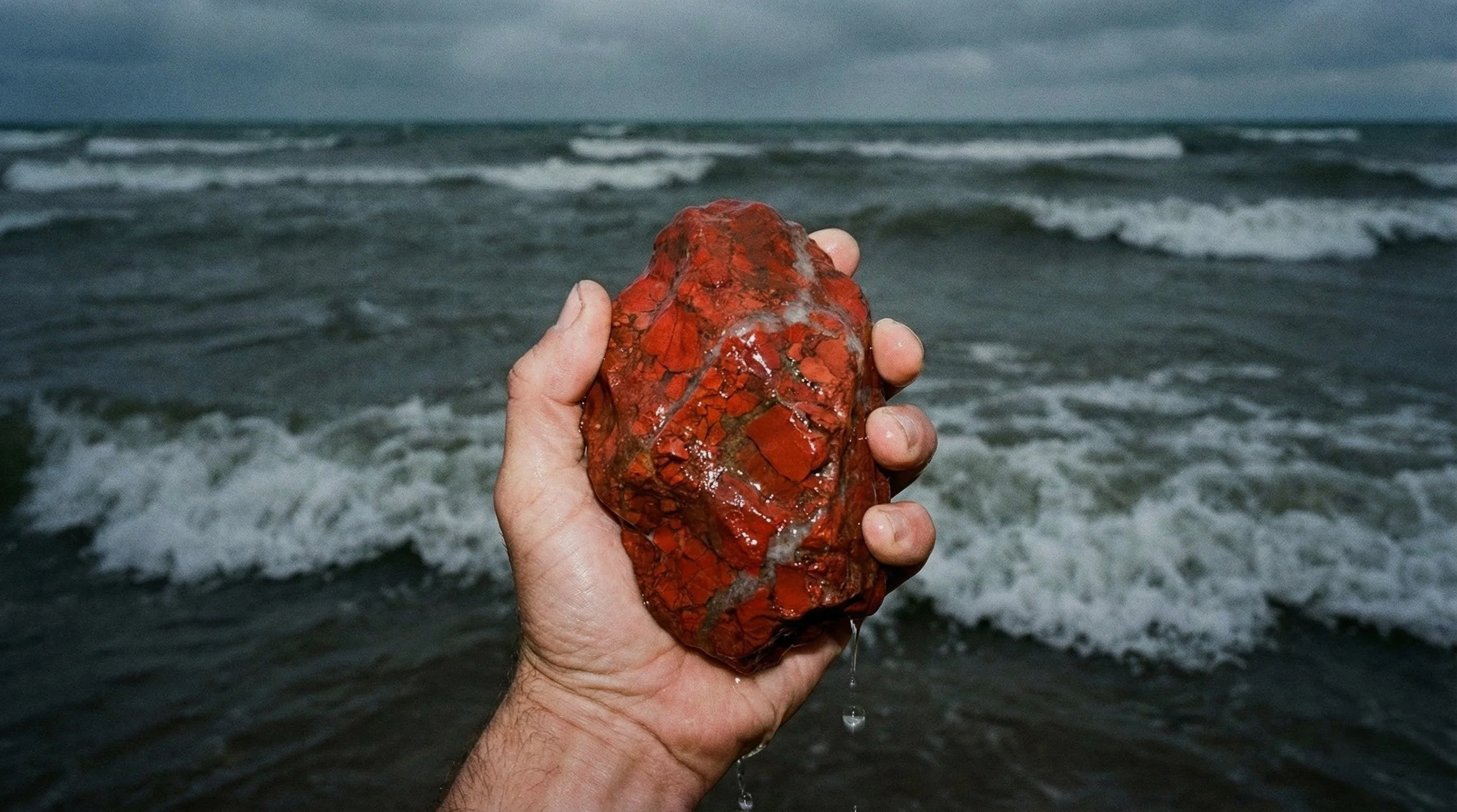 A person's hand holding a large red-orange rock with a wet surface, against a backdrop of a cloudy sky and ocean waves.