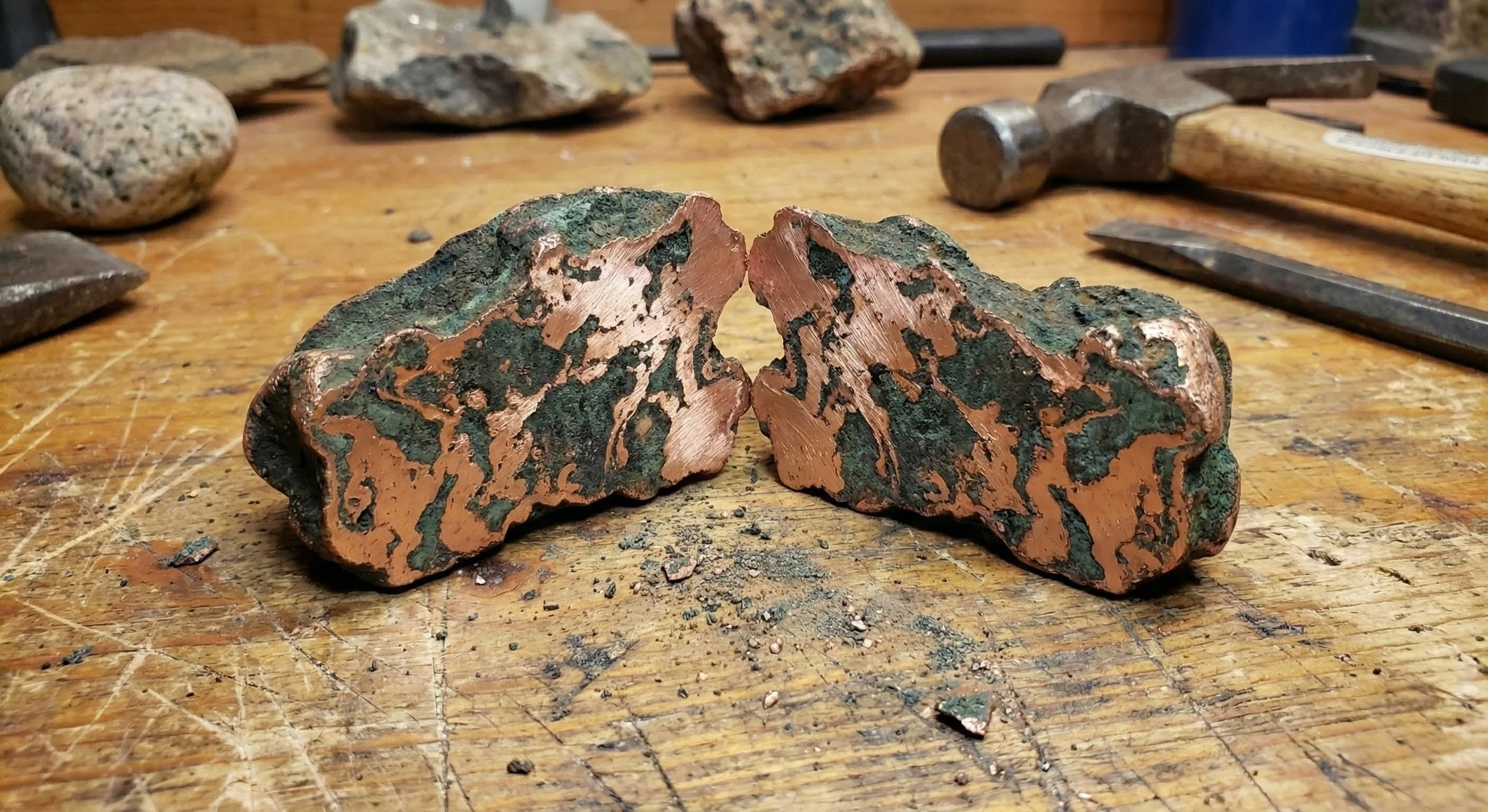 Two broken pieces of copper ore with green patina and reddish-brown inner surface, placed on a wooden workbench surrounded by tools and additional rocks.