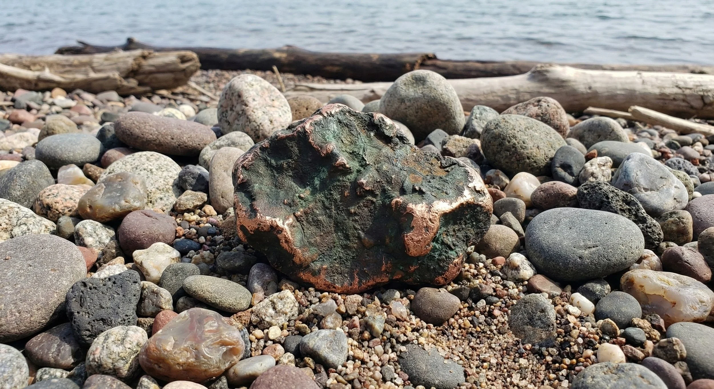 Close-up of a large, rugged, greenish-brown rock on a pebble-covered shoreline with driftwood and water in the background.