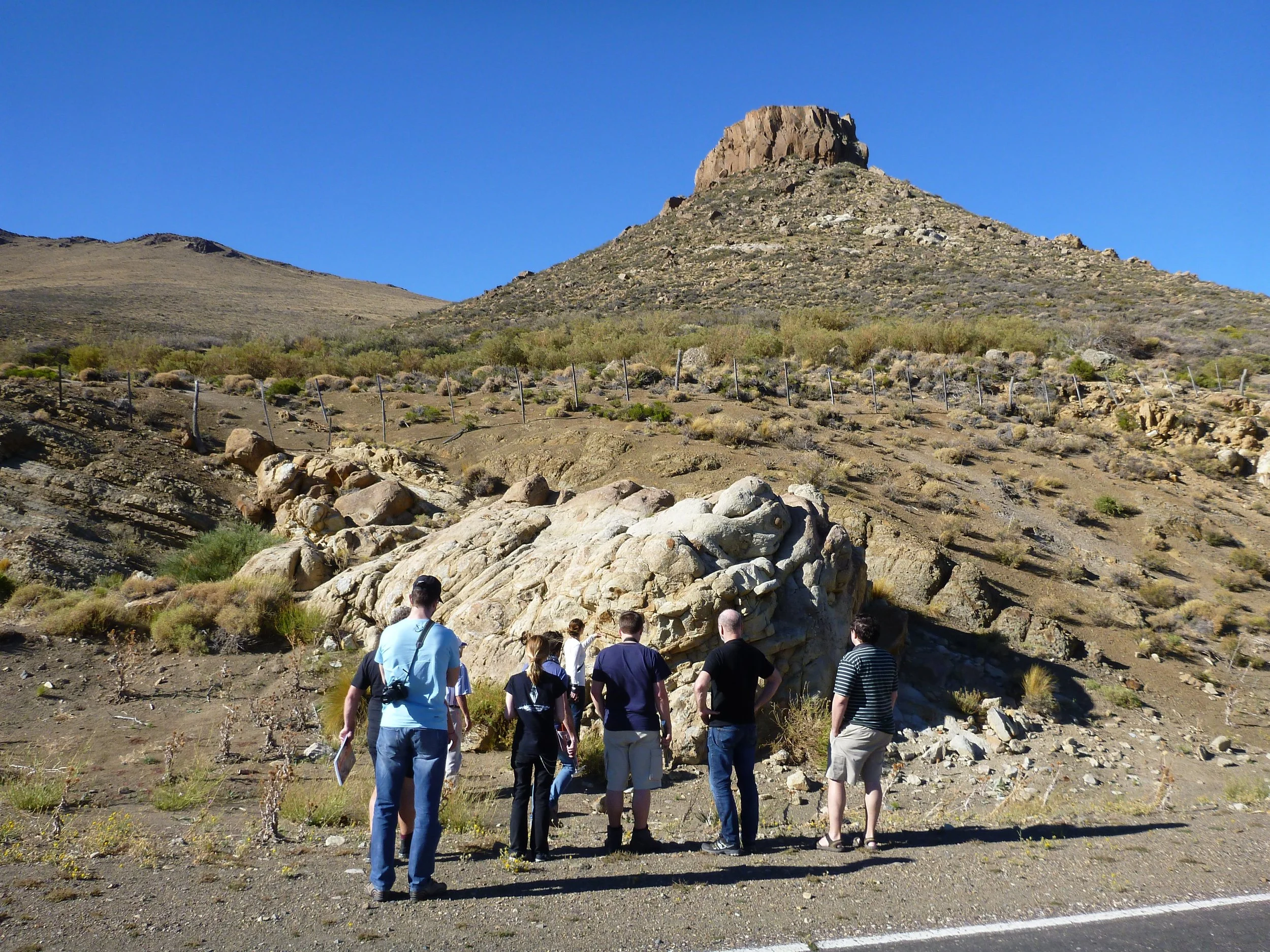 A group of people gathered outdoors in a desert landscape with rocks and sparse vegetation, looking at a large rock formation under a clear blue sky.