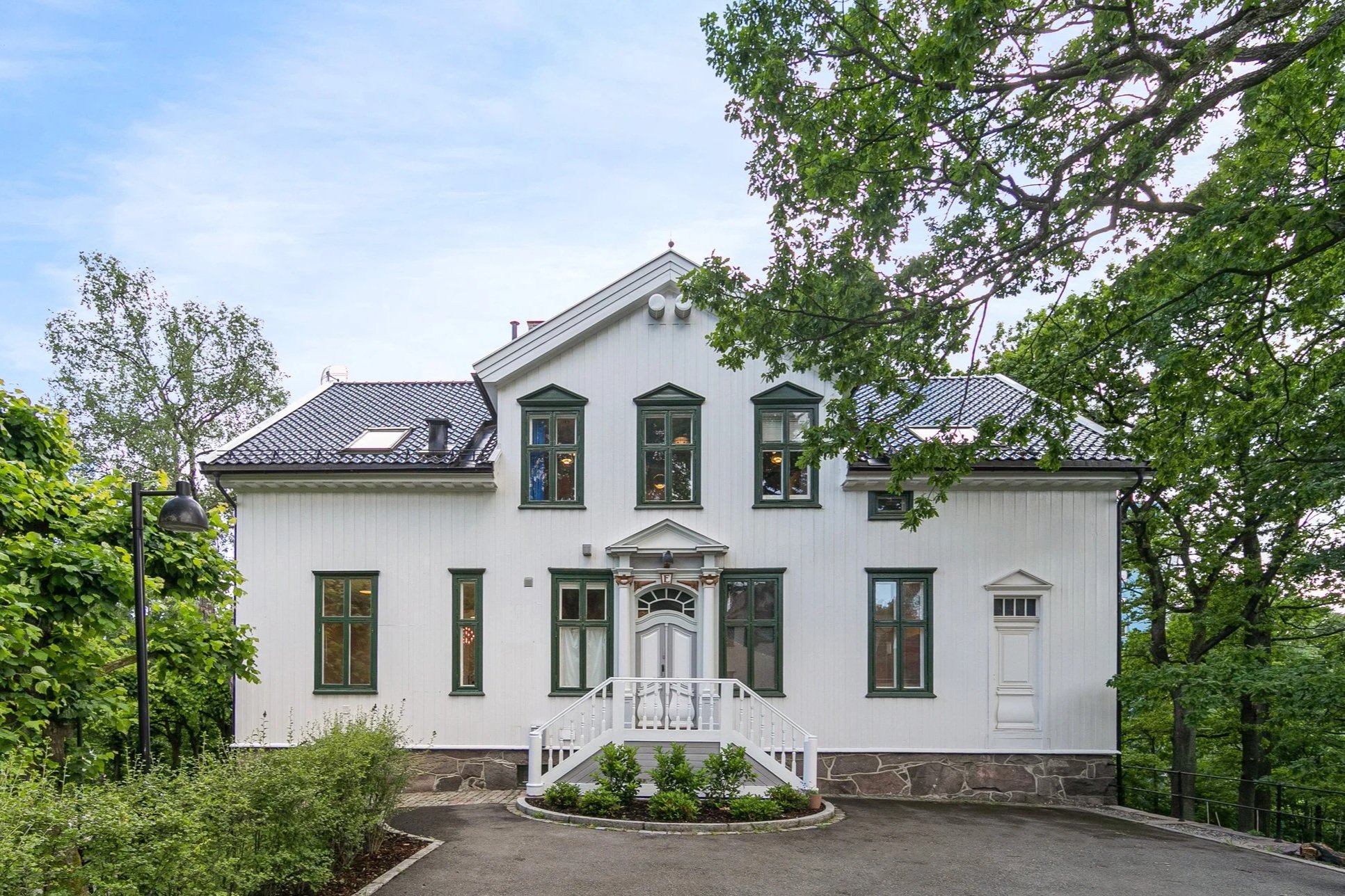 White house with dark green trim, surrounded by trees, a small set of stairs with white railing leading to the front door, and a landscaped garden in front.