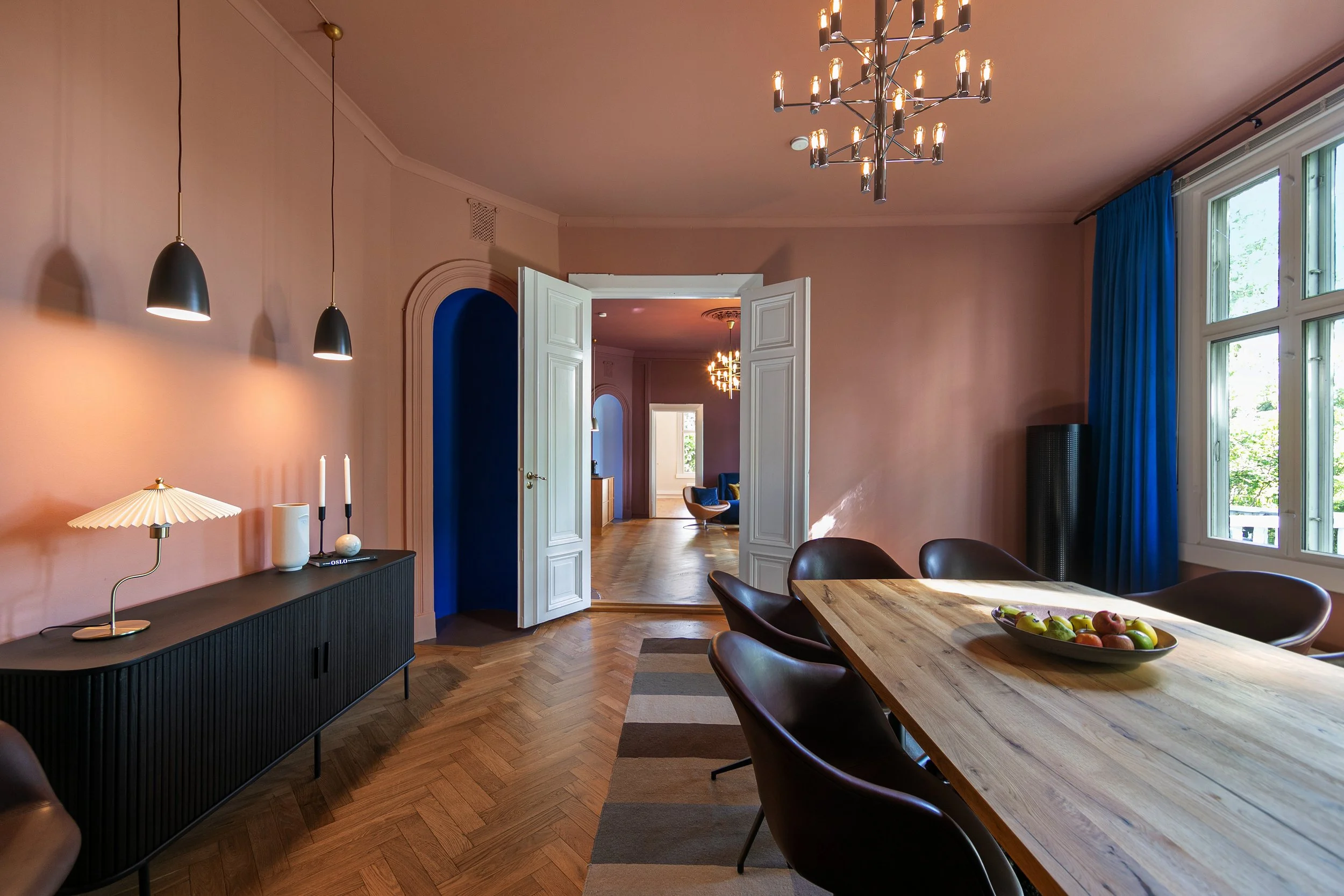 A dining room with a wooden table, six black chairs, a black sideboard, a bowl of fruit, large windows with blue curtains, pendant lights, and a chandelier.