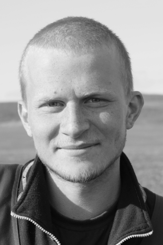 Black and white close-up portrait of a young man with short hair outdoors, wearing a zip-up jacket.
