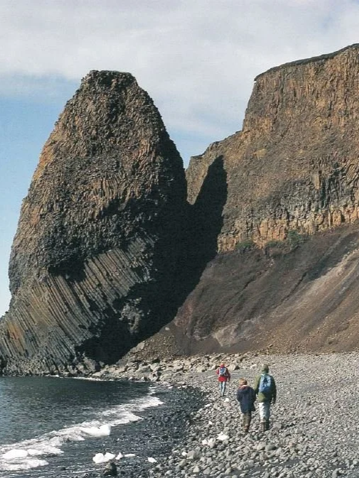 Three hikers walking along a rocky beach with large cliffs and a sea stack in the background.