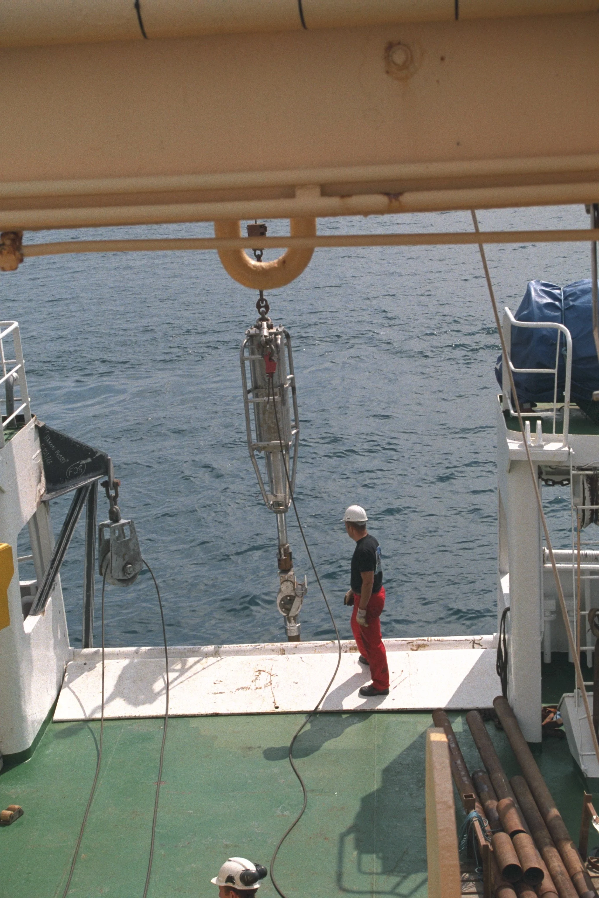 A man on a ship's deck near the ocean, wearing a white helmet, a black t-shirt, and red pants, standing next to a large submersible or underwater vehicle hanging from the ship's crane.