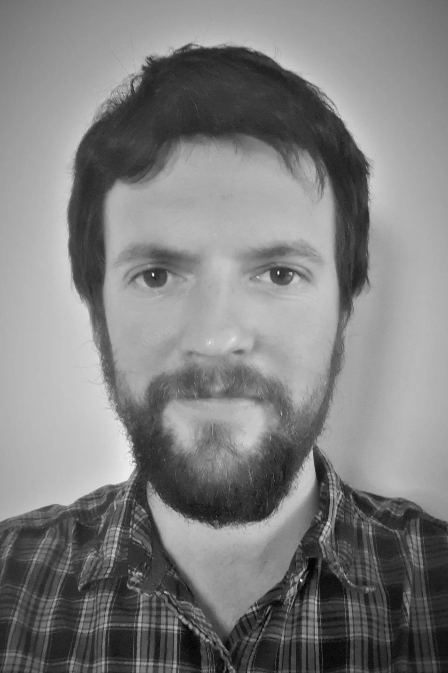 A black and white photo of a man with dark hair and a beard, wearing a plaid shirt, looking directly at the camera with a neutral expression.