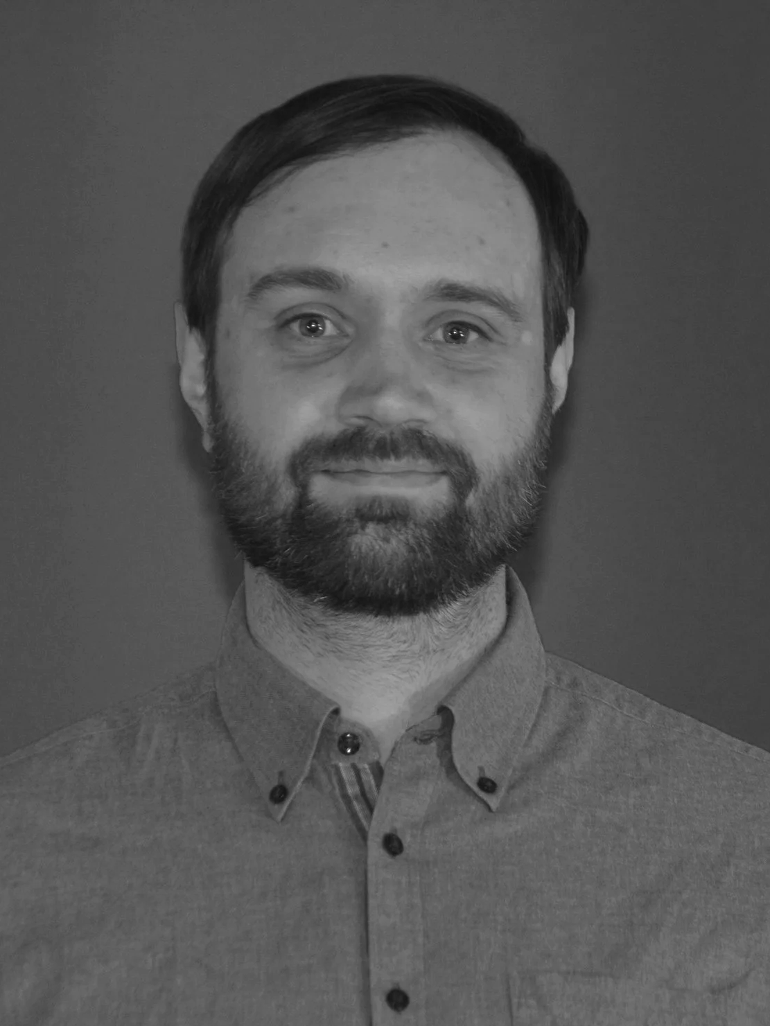 A black-and-white portrait of a man with short dark hair, a beard, and a mustache, wearing a button-up shirt, looking directly at the camera with a slight smile.