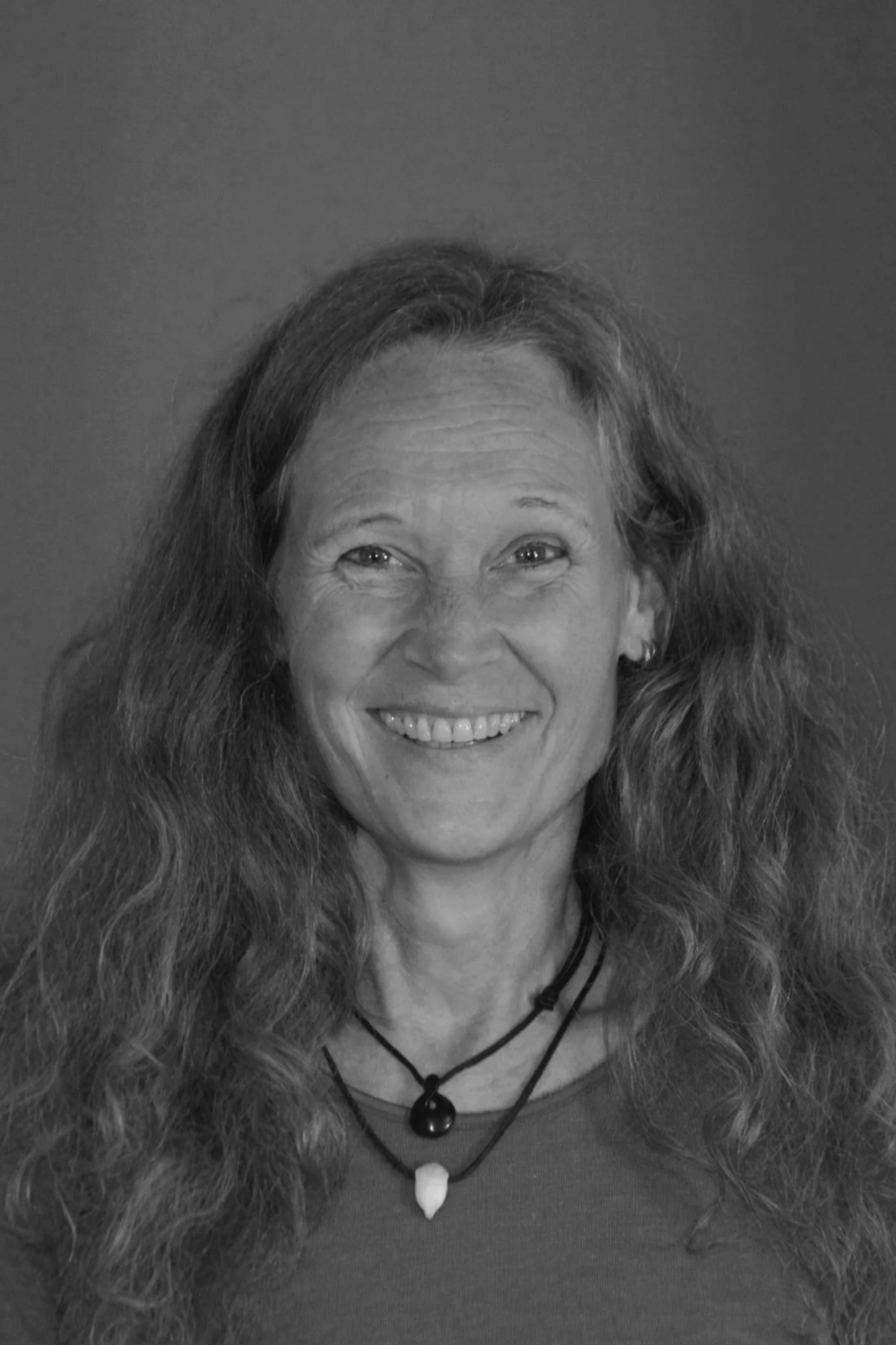 A smiling woman with long curly hair and earrings, wearing layered necklaces, looking at the camera against a plain background.