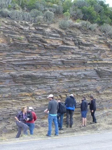 Group of six people studying a rock formation on a hillside beside a road.
