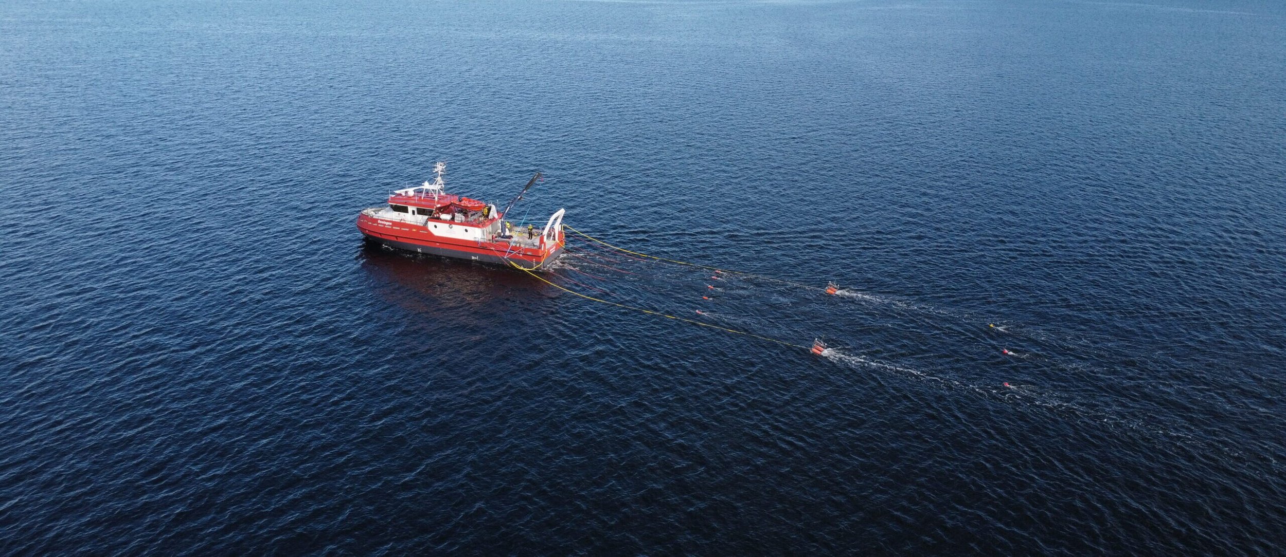A red and white research vessel in open water, deploying equipment with long yellow cables behind it.