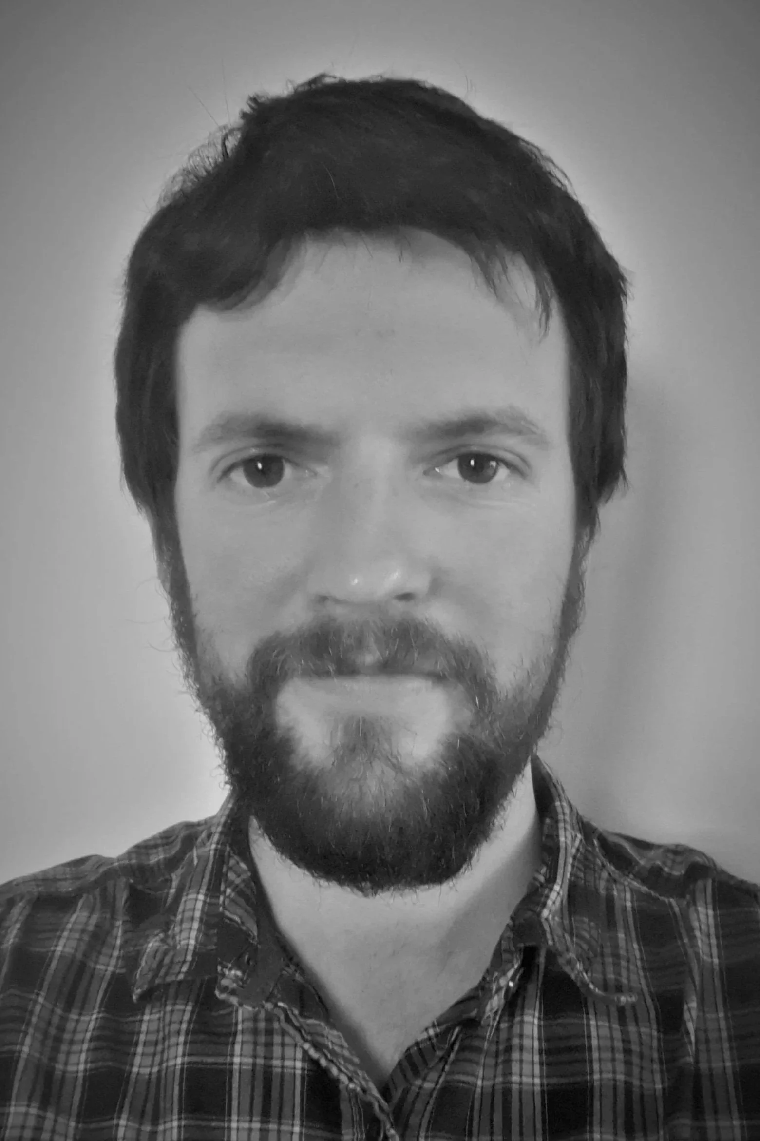 Black and white photo of a man with short dark hair, a beard, and mustache, wearing a plaid shirt, looking directly at the camera.