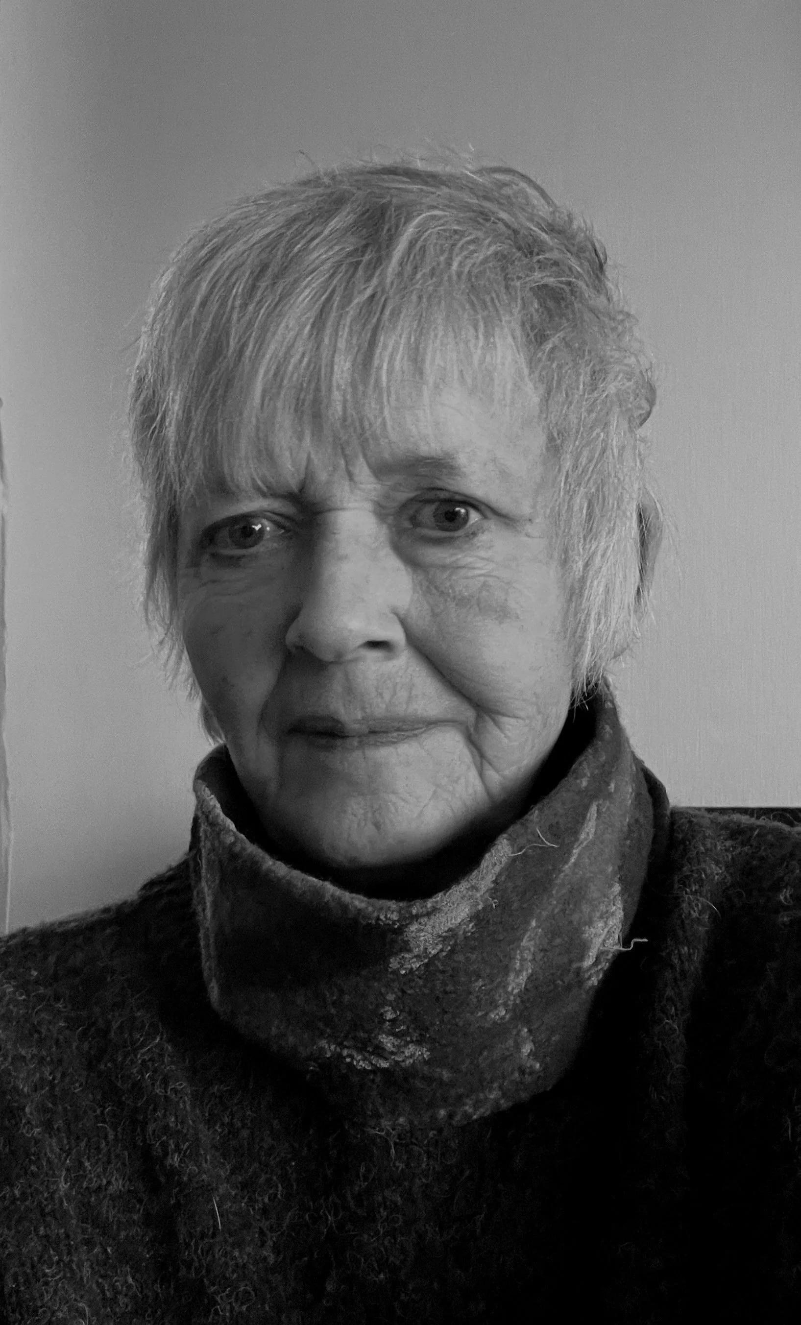 Black and white photo of an elderly woman with short hair, wearing a scarf and a sweater, looking into the camera with a neutral expression.