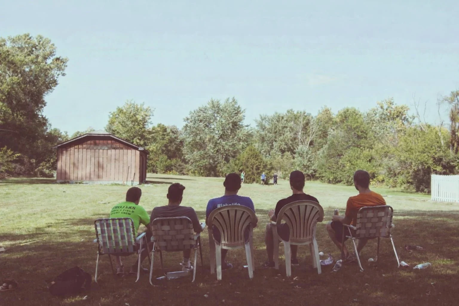 Five men bonding on lawn chairs face a grassy yard and trees, with a brown shed visible on the left.