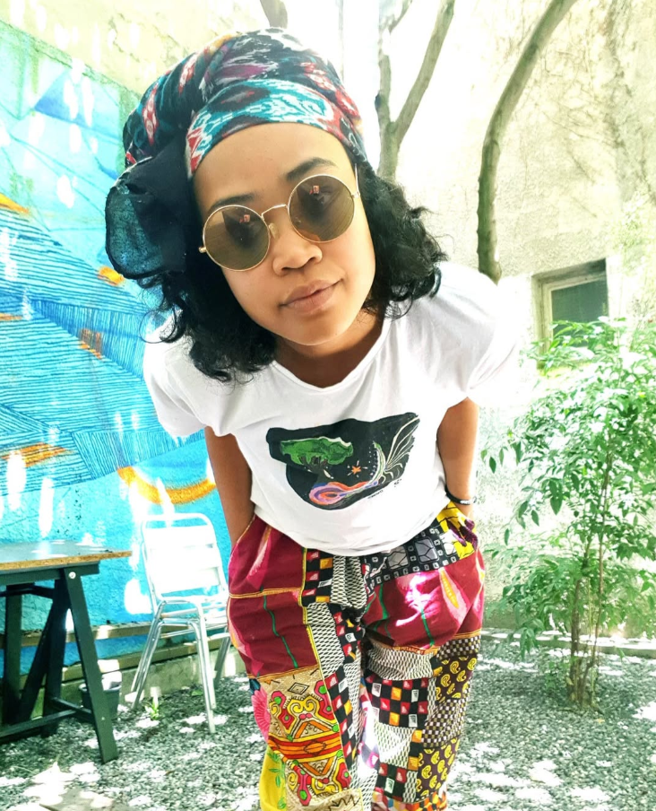 Femme portant des lunettes rondes, un bandana coloré, un t-shirt blanc et un pantalon vibrant avec différents motifs, debout à l'extérieur avec des arbres et une fresque murale en arrière-plan.