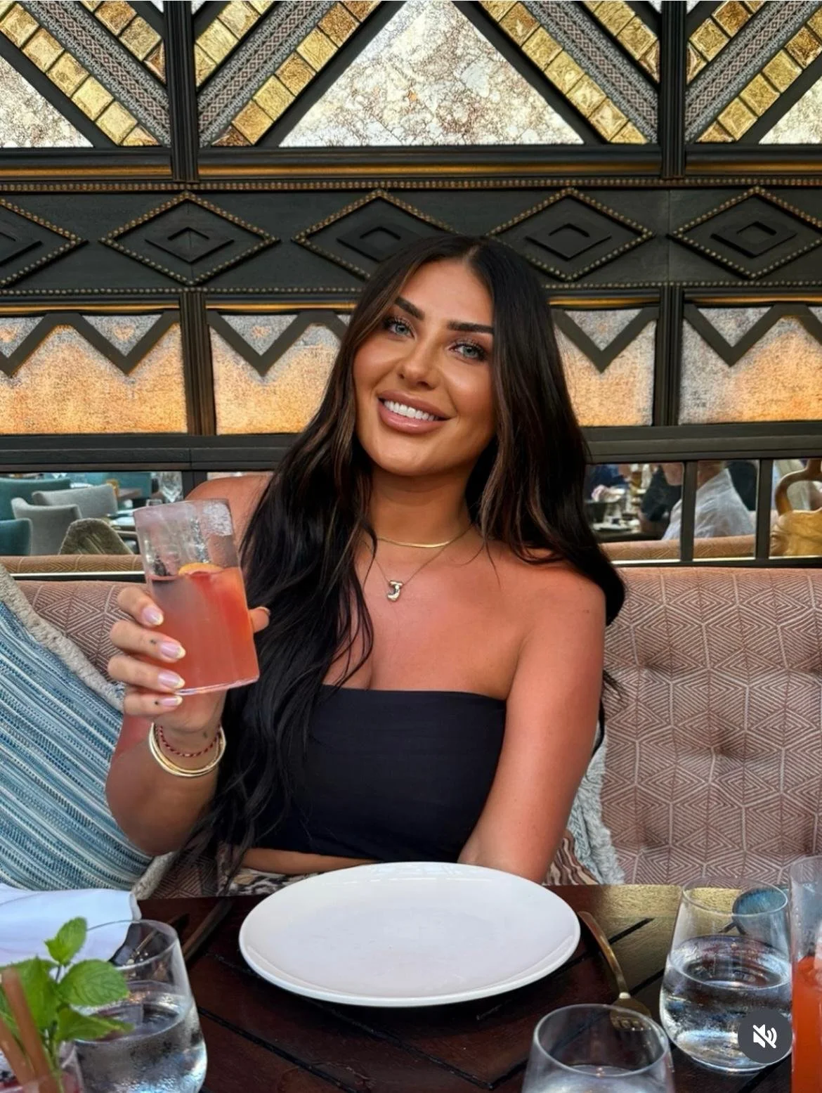 A woman seated at a restaurant table holding a glass of pink drink, smiling at the camera, with tableware and glasses on the table, and a decorative wall in the background.