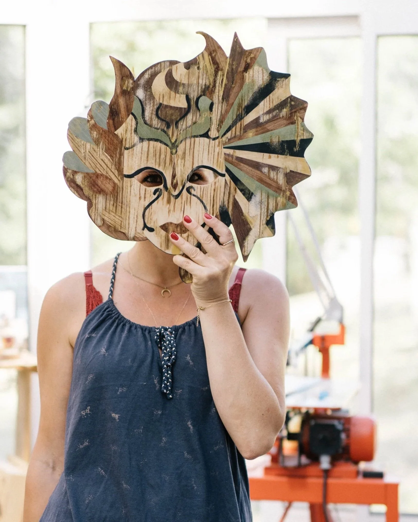 Une femme porte un masque en forme de lion en bois et pose devant une machine d'atelier.