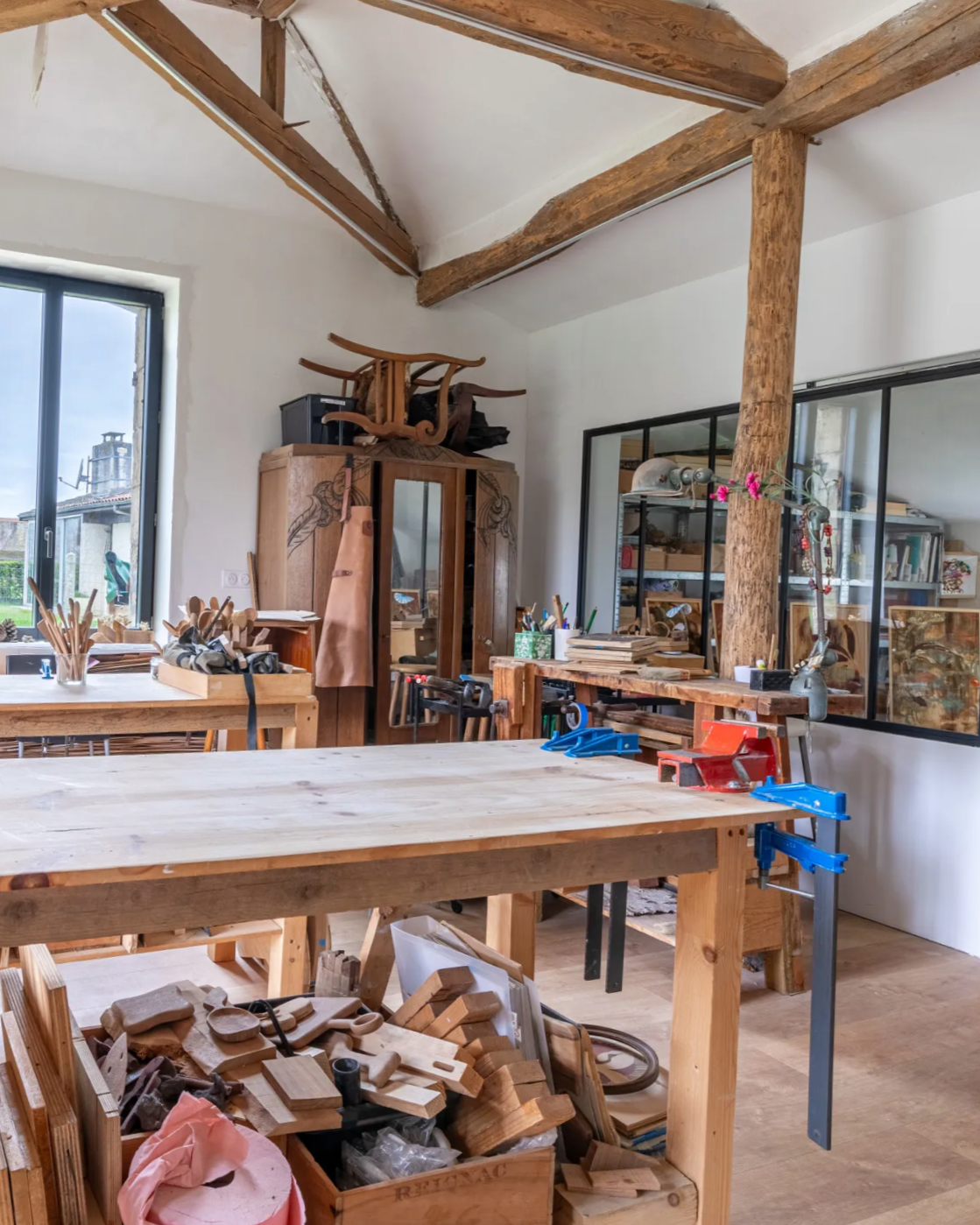 Atelier de menuiserie avec tables en bois, outils, matériaux et meubles en bois, grande fenêtre laissant entrer la lumière naturelle, poutres en bois au plafond.