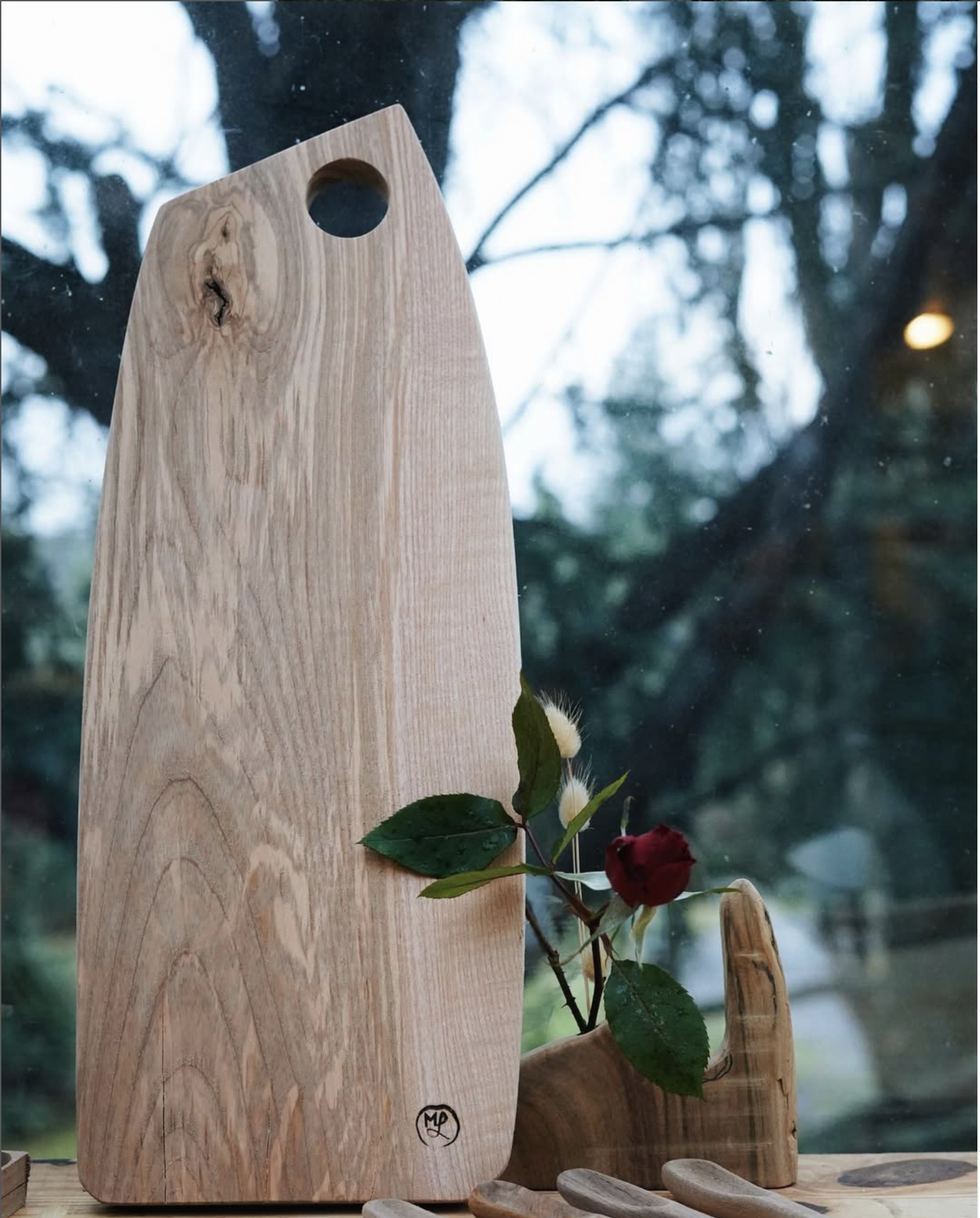 Suppoort en bois avec une rose rouge et des feuilles vertes, posé sur une surface en bois, avec une fenêtre en arrière-plan montrant des branches d'arbres et un ciel nuageux.