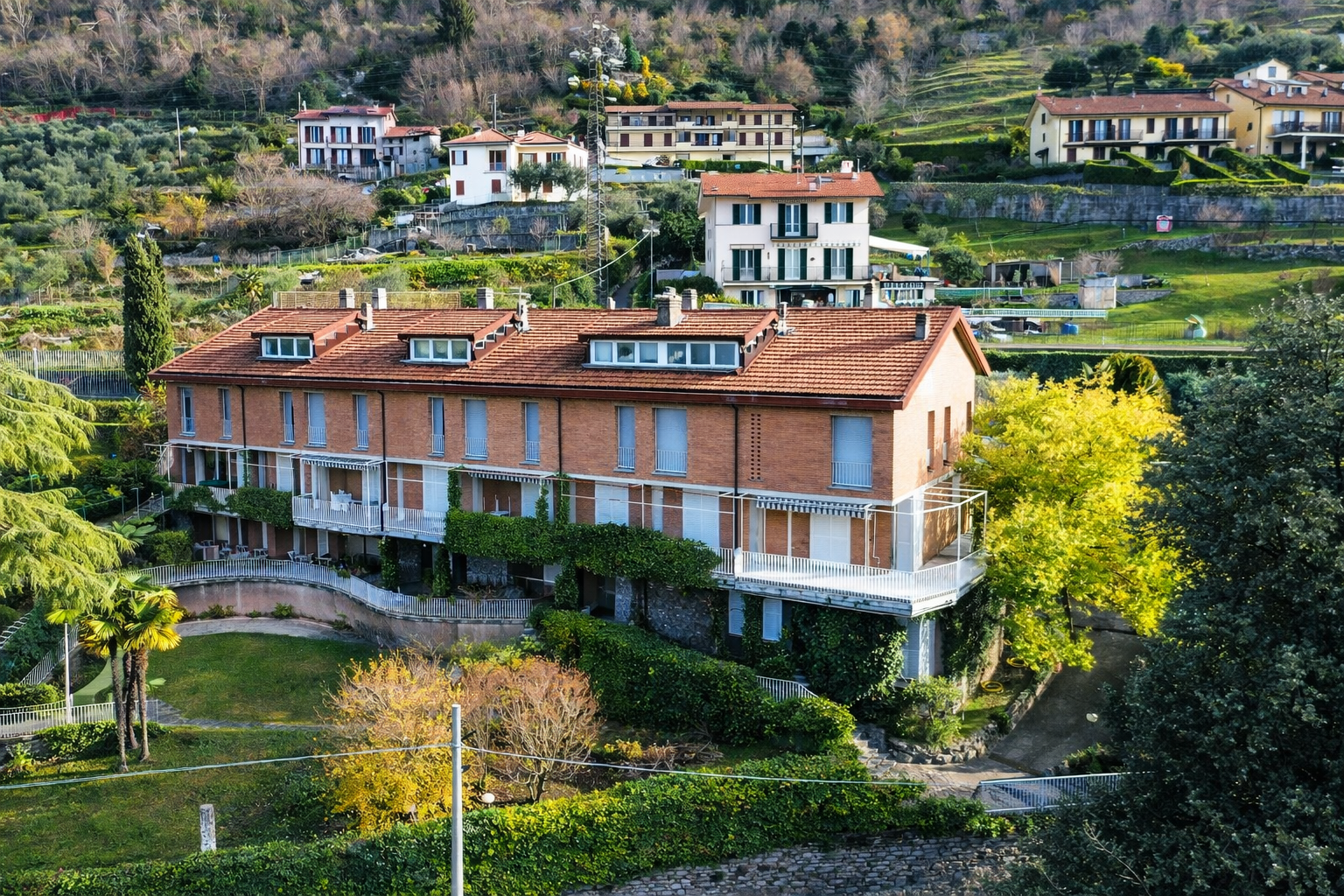Un villino di tre piani con tetto rosso e terrazze, circondato da giardini verdi, in una zona collinare con altre case e alberi.
