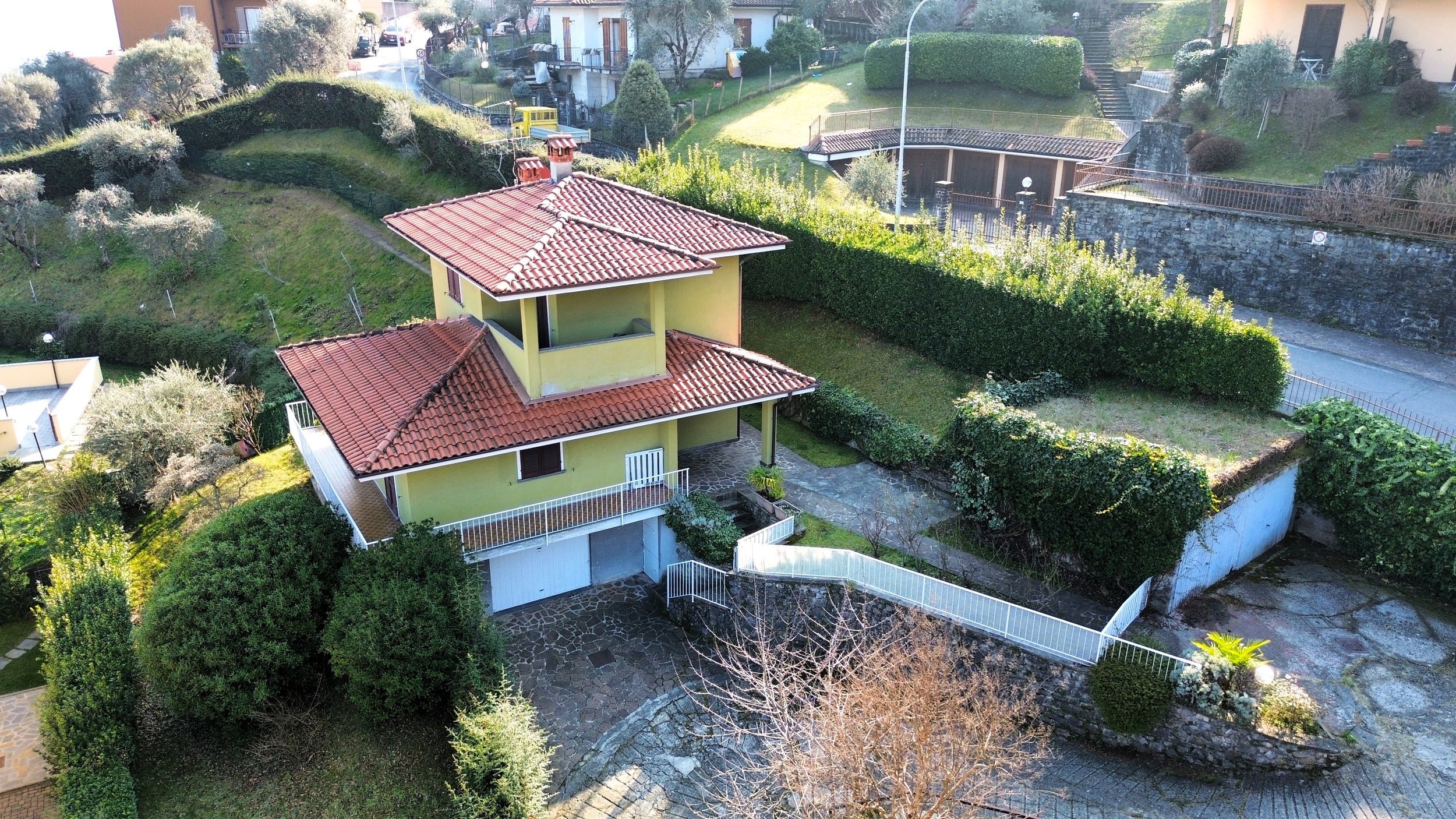 Casa gialla con tetto rosso, situata in una zona collinare circondata da cespugli e alberi, con vialetti e recinzioni bianche, e un terreno cortilizio in pietra.