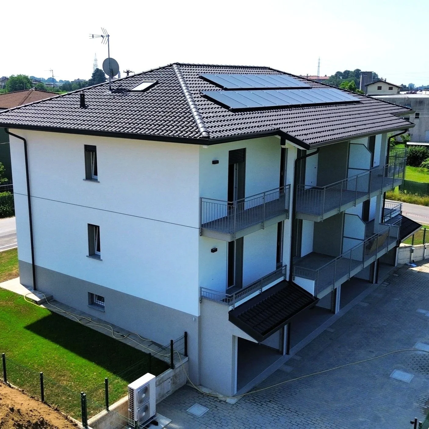 Edificio residenziale con terrazze, tetto con pannelli solari e antenne, circondato da un giardino e recinzione.
