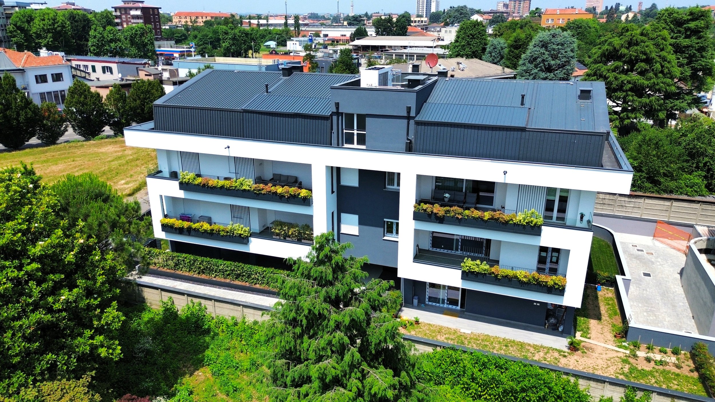 Immagine di un edificio moderno con balconi e piante, circondato da verde, in una zona urbana con altri edifici e alberi.