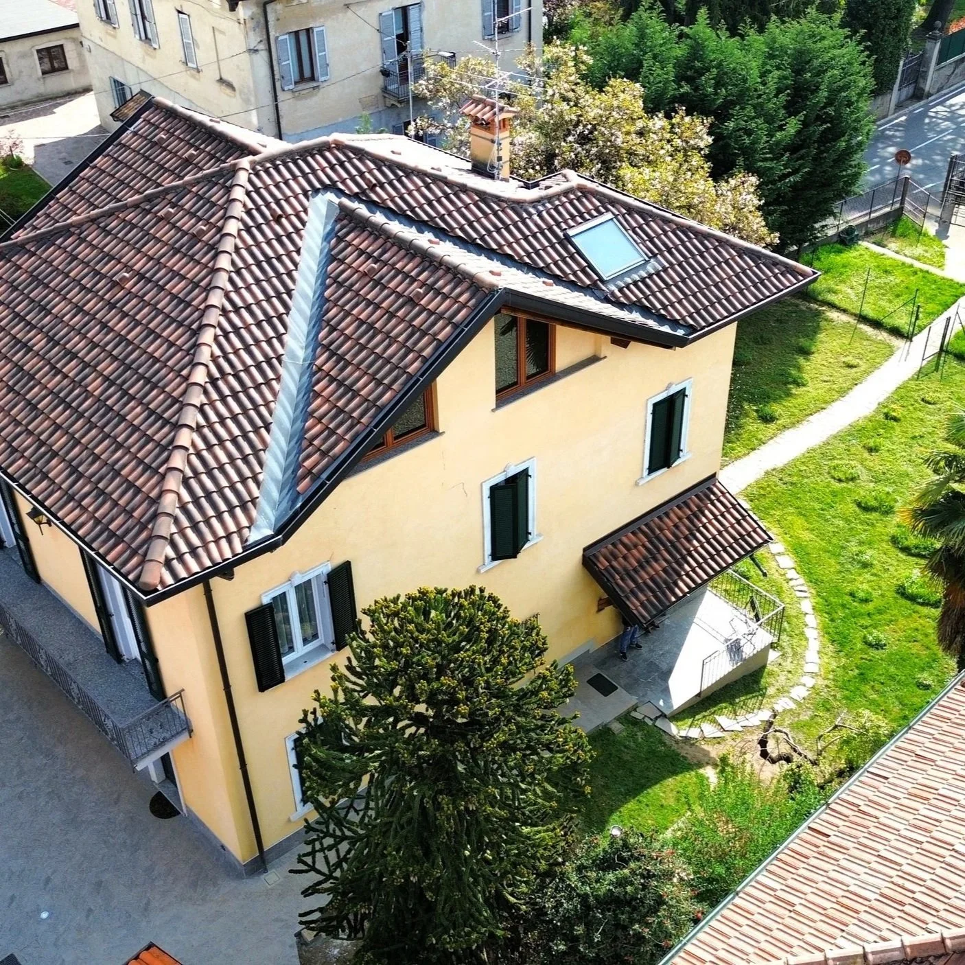 Vista aerea di una casa con tetto in terracotta e pareti gialle, circondata da un giardino verde con alberi e un percorso di pietre.