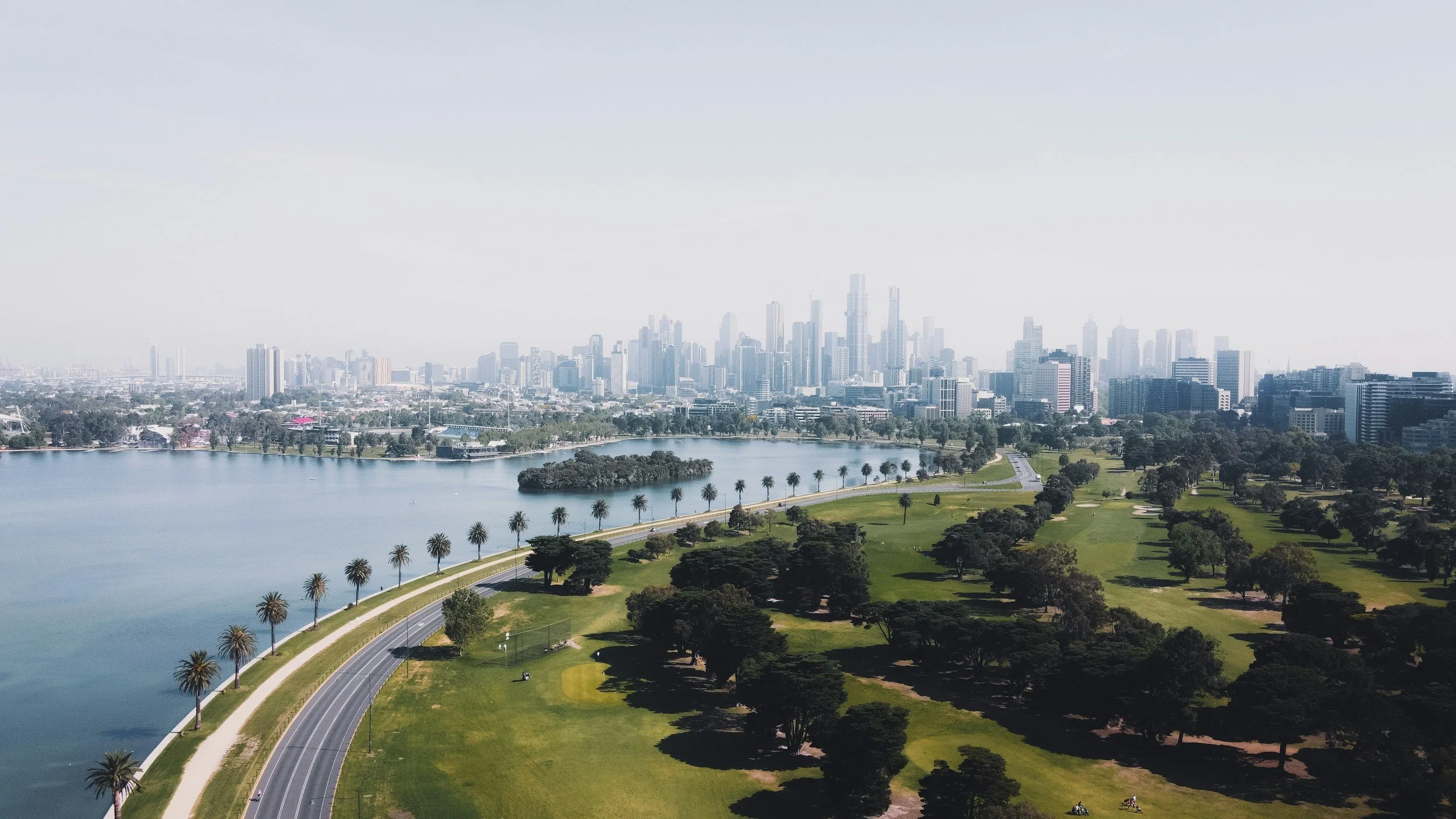 Aerial view of a city skyline with tall skyscrapers, a lake with a tree-lined shoreline, and a park with green grass, trees, and a curved road.