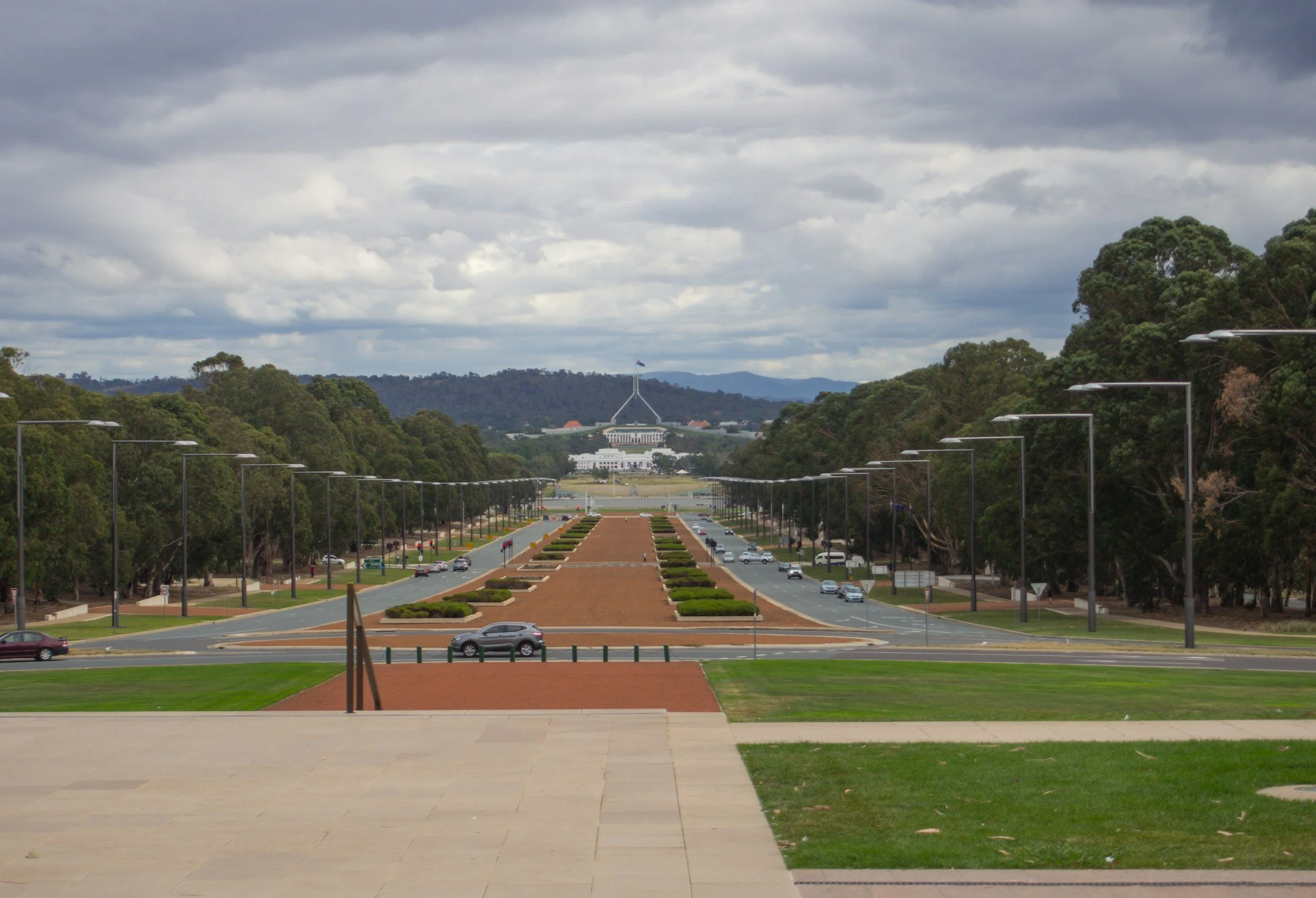 The image depicts the view from the steps of the Australian Parliament House, overlooking a long, symmetrical driveway lined with streetlights, greenery, and cars, with the Parliament House building and the city skyline in the distance under a cloudy sky.