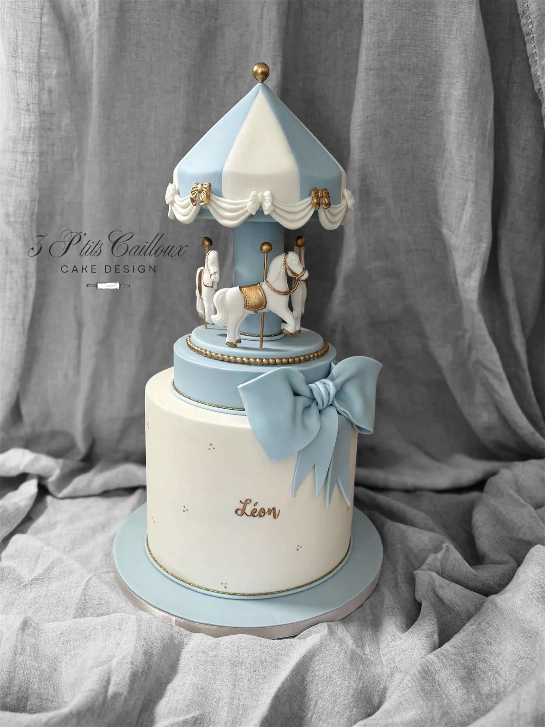 Gâteau en forme de carrousel de cheval, décoré de blanc, bleu, or et avec une grande nœud bleu, avec le nom 'Léon' en or sur la base.
