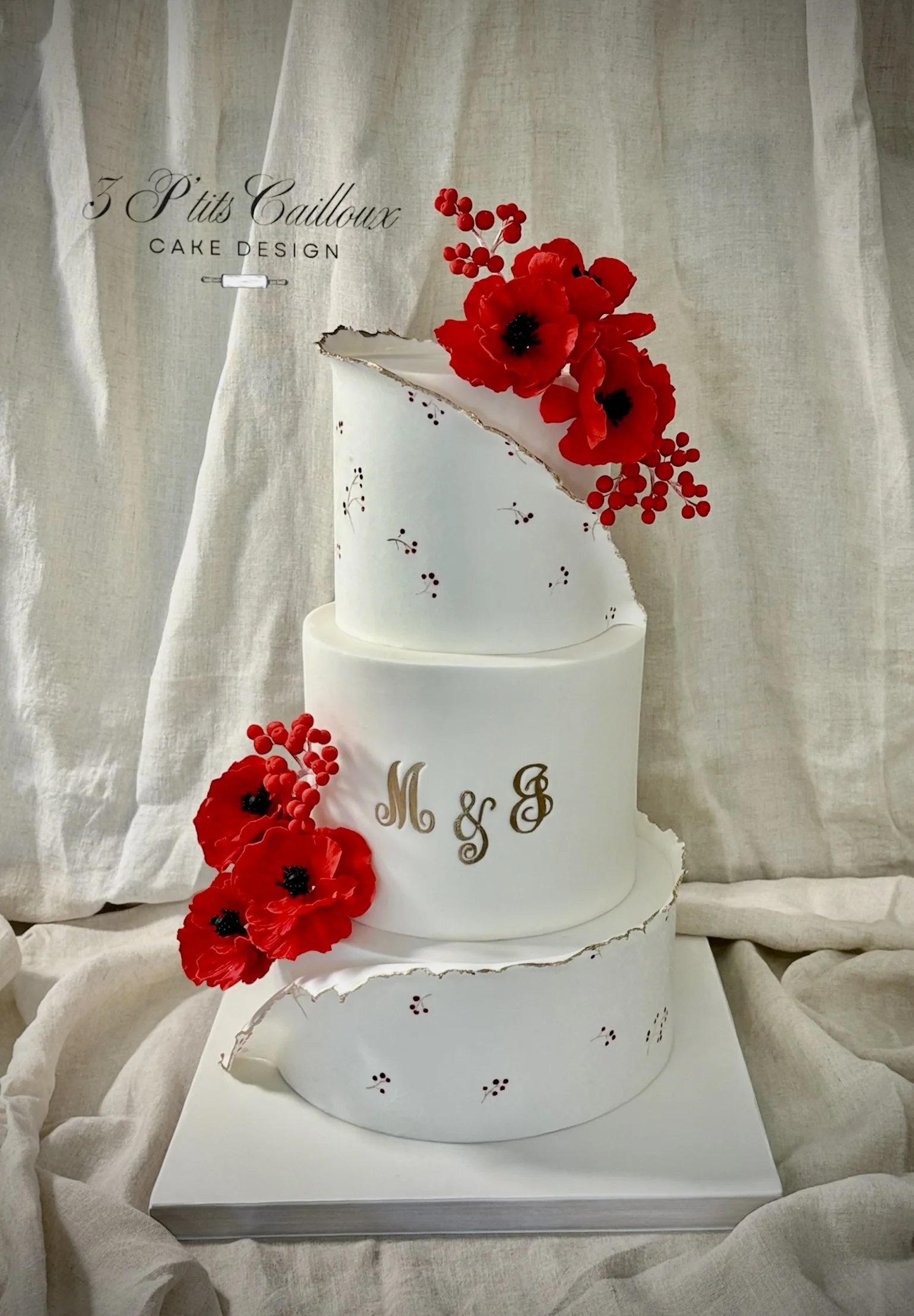 Un gâteau blanc à trois niveaux orné de fleurs rouges artificielles, avec des lettres dorées M & G sur le niveau du milieu, et une décoration en céramique imitant une fissure en or, présenté sur un support blanc devant un rideau beige.