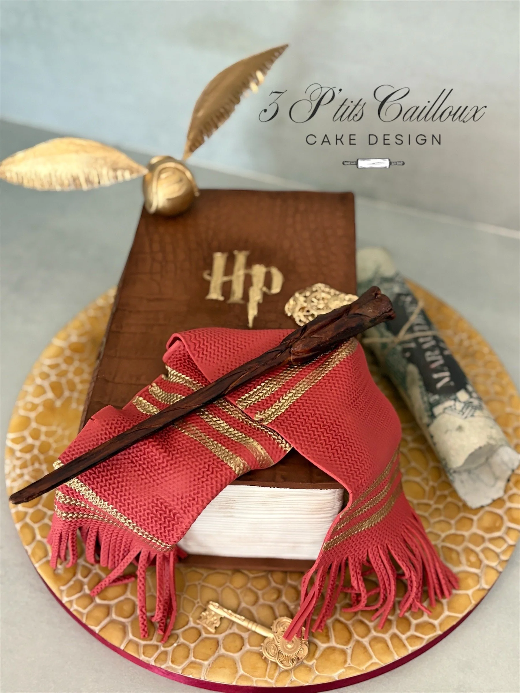 Gâteau en forme de livre avec une écharpe rose, une baguette en bois, un rouleau de parchemin et des décorations dorées sur un plateau ronds.