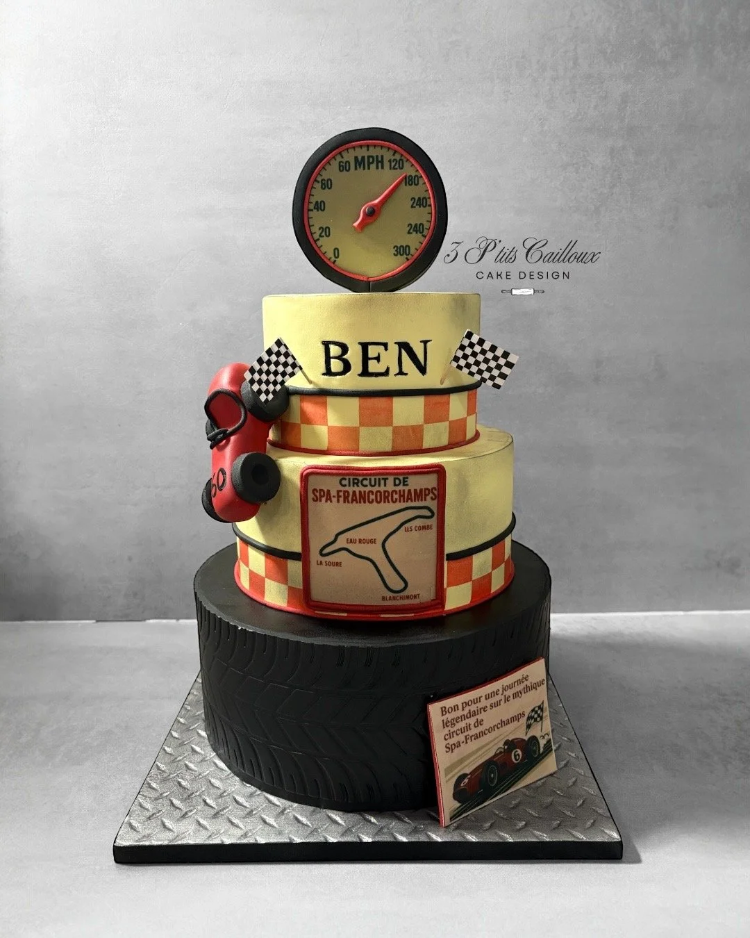 Gâteau en forme de circuit de course avec un pneu comme base, décoré de couleurs jaunes et rouges, avec une petite voiture de course sur une des couches, un tableau avec un circuit, un panneau disant "BEN" et un compteur de vitesse affichant 180 MPH.