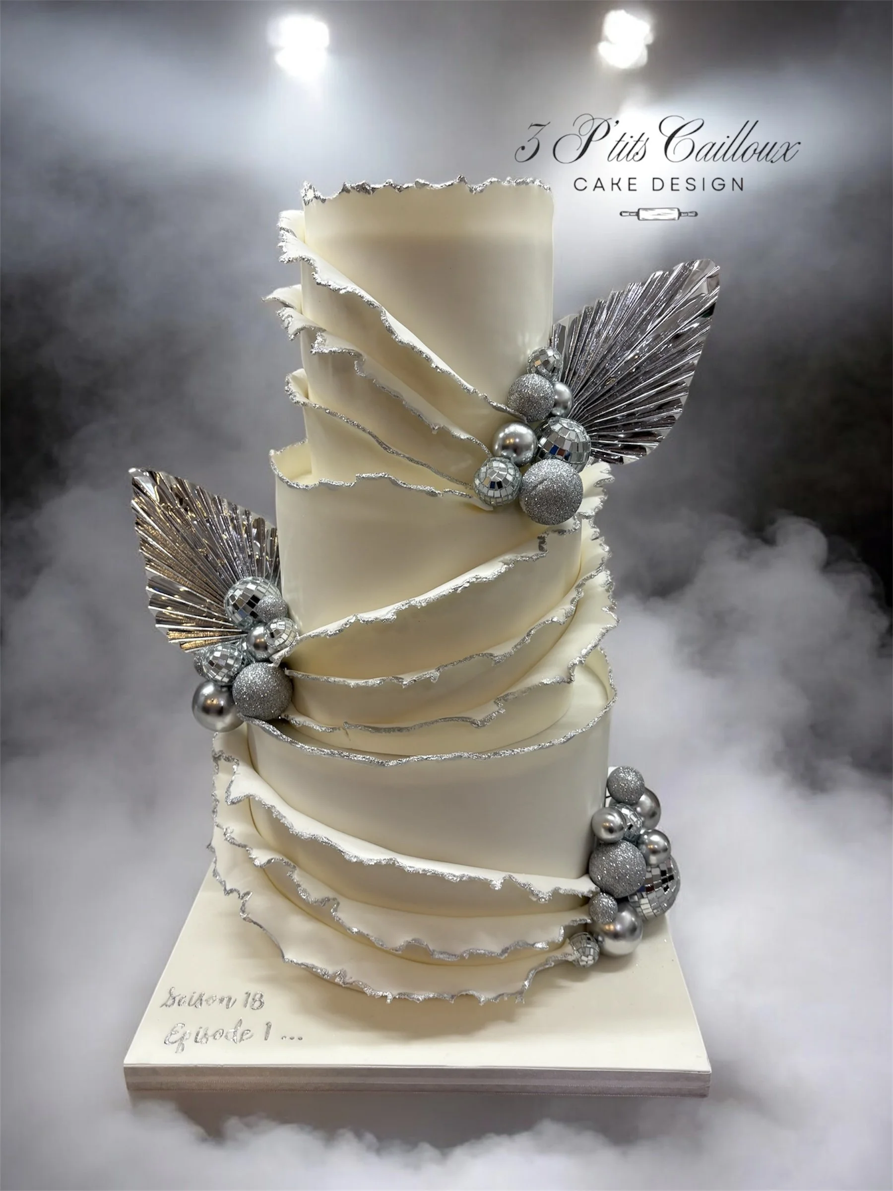 Gâteau blanc en forme de spirale avec décorations argentées, feuilles en aluminium, boules argentées et paillettes, présenté sur une base blanche avec des nuages de fumée en arrière-plan.