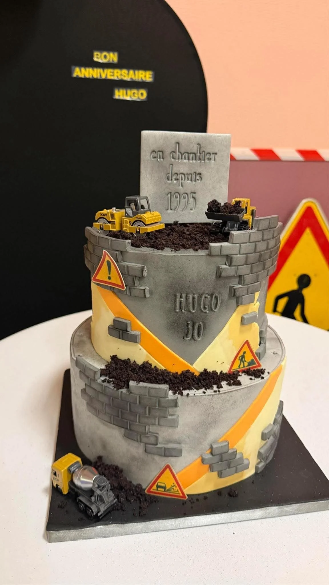 Gâteau d'anniversaire pour Hugo, décoration de construction avec des petits camions, des panneaux de signalisation, des briques en biscuit, et un message en pierre indique 'en chantier depuis 1995'.