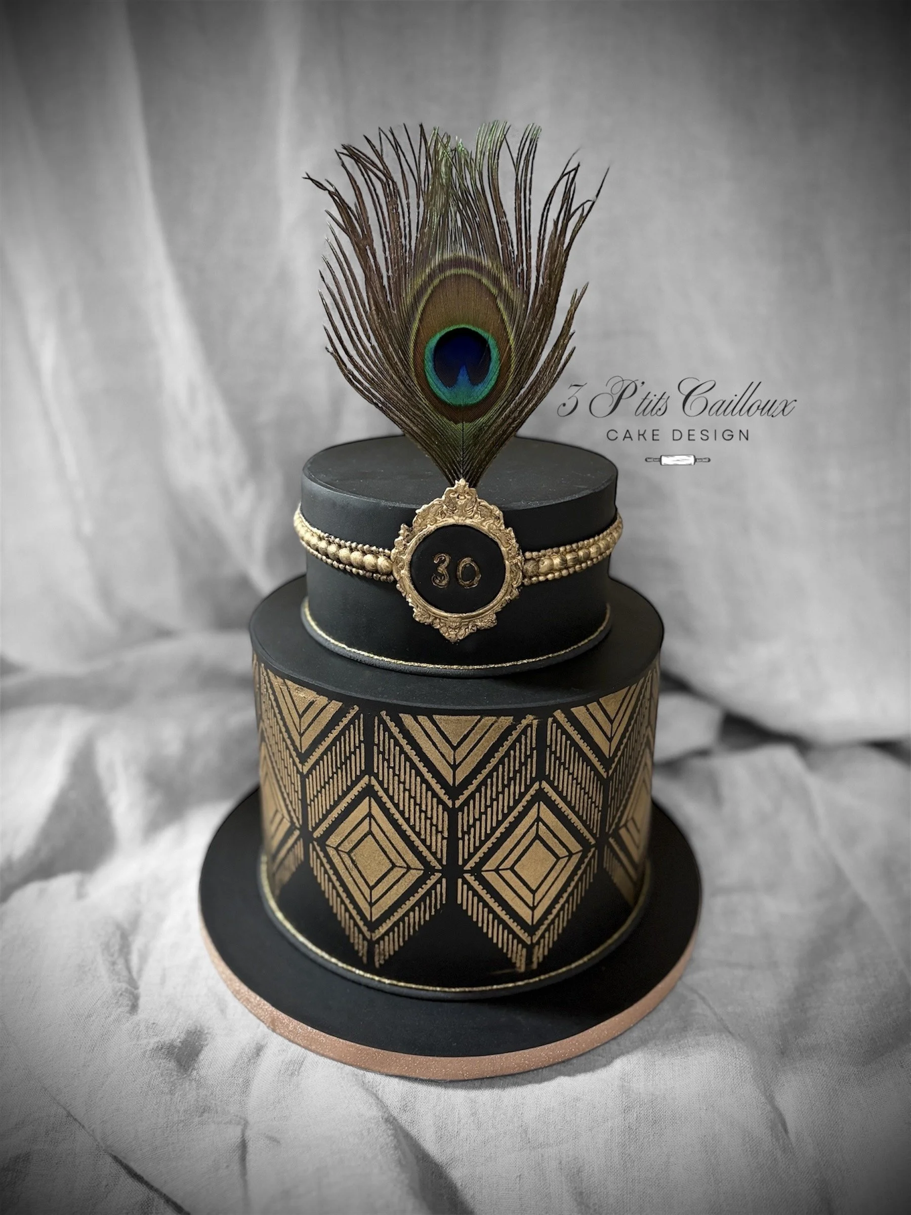 Gâteau deux étages avec décoration noire et or, motif géométrique sur la base, plume de paon en haut, badge doré avec le nombre 30, écrit en français "3 Petits Cailloux" et "Cake Design".