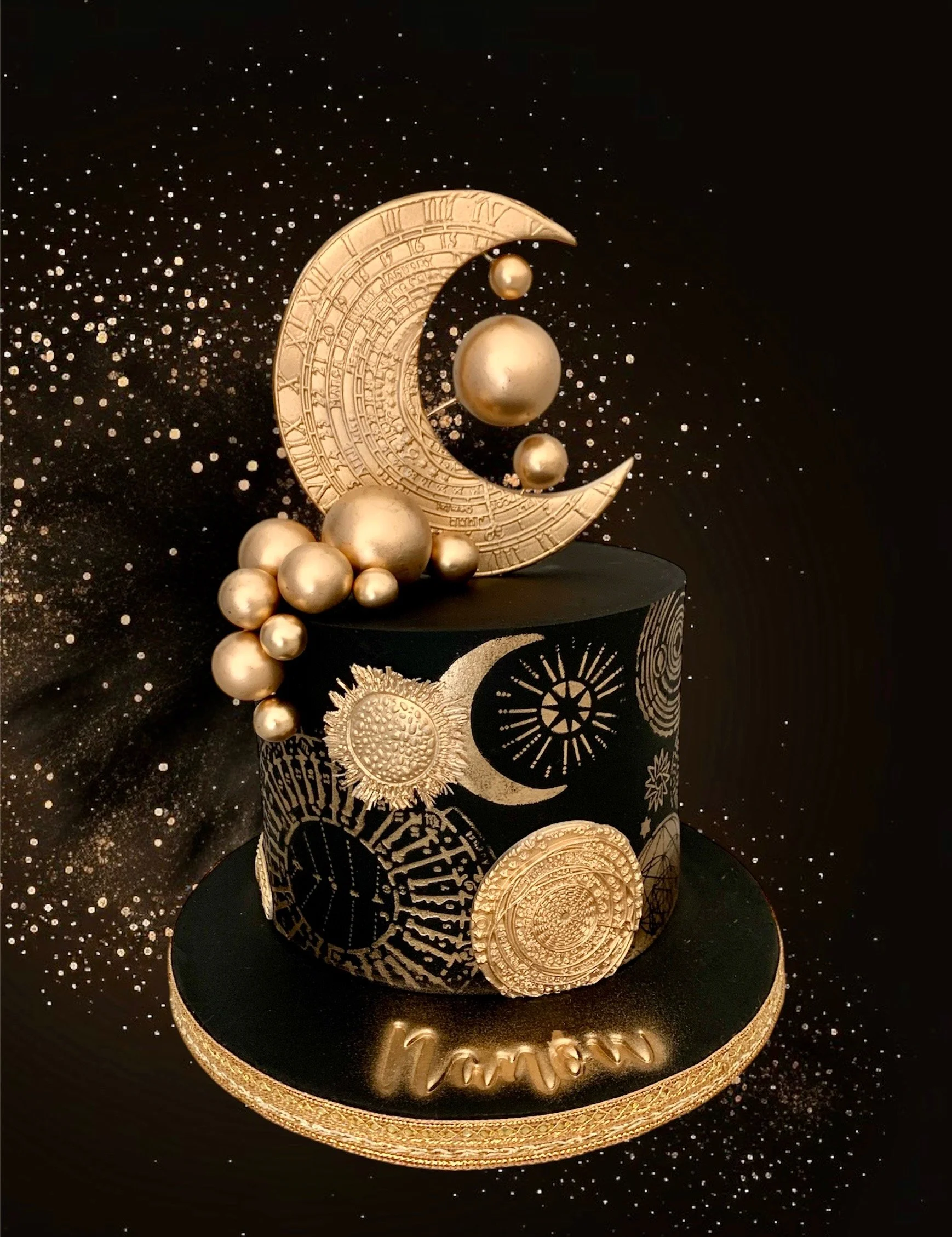 Gâteau noir décoré de motifs dorés, avec un décor de lune, de planètes et de symboles cosmiques, et des sphères dorées, avec une inscription "Nour" en or sur le socle.