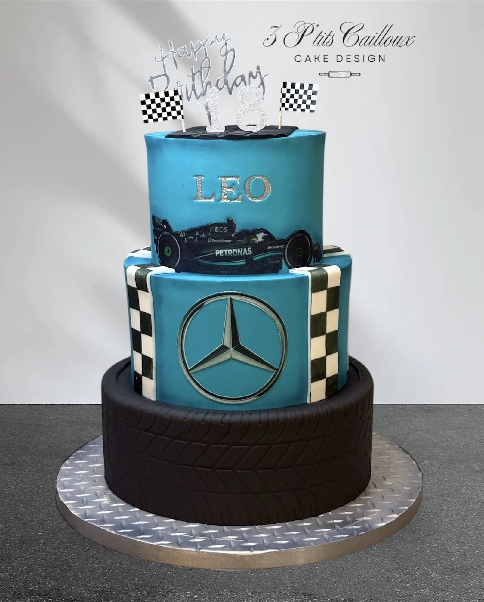 Gâteau d'anniversaire à plusieurs niveaux avec des thèmes de voitures de course et de Mercedes, décoré avec un symbole Mercedes, un logo Renault, une voiture de Formule 1, et un message de fête avec un topper "Happy Birthday" et un chiffre 18.