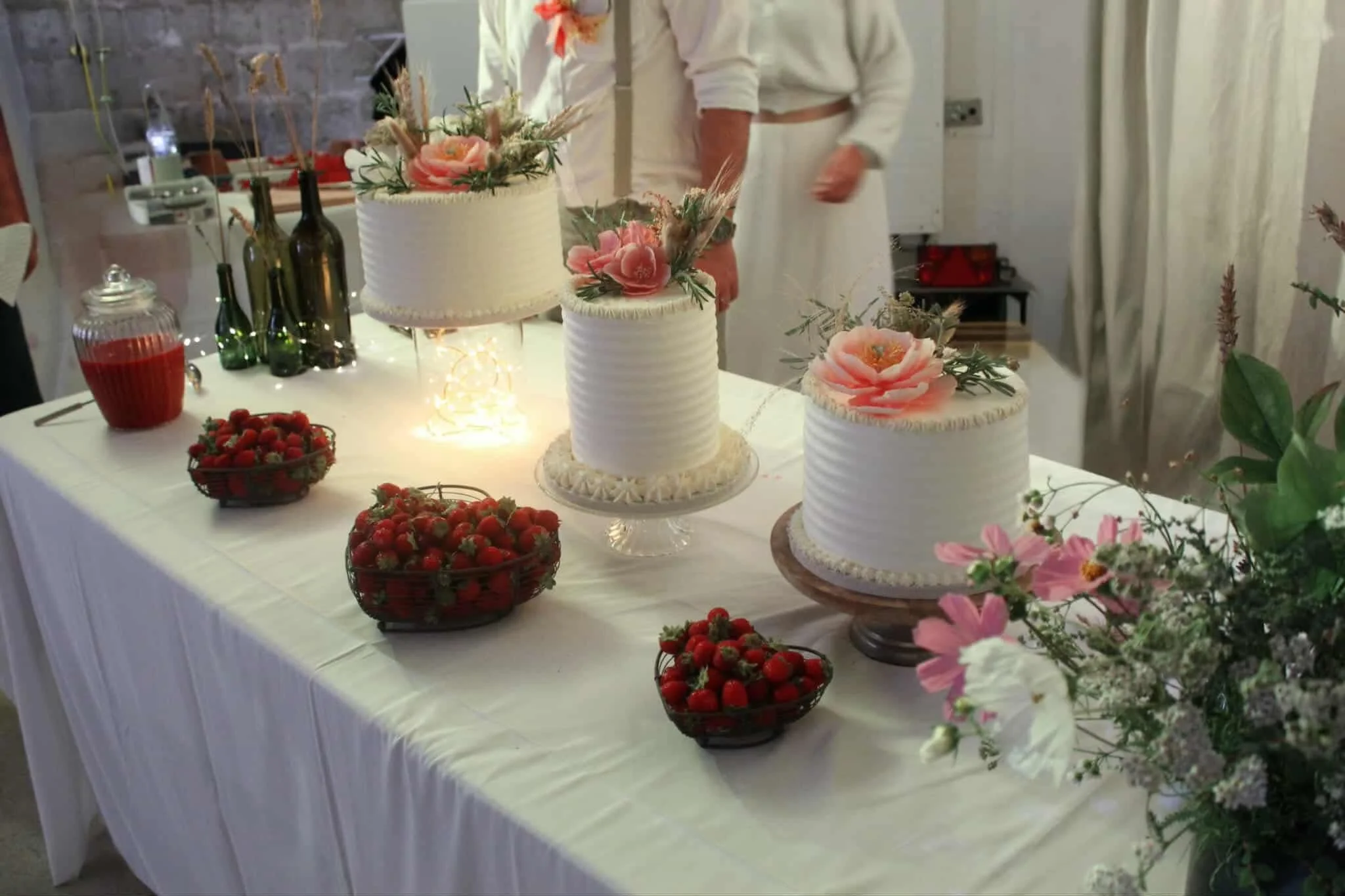 Trois gâteaux roses décorés de fleurs roses et de feuillage, sur une table blanche avec des paniers de fraises. Il y a des bouteilles et des bocaux en verre derrière les gâteaux, ainsi que des personnes en vêtement blanc en arrière-plan.