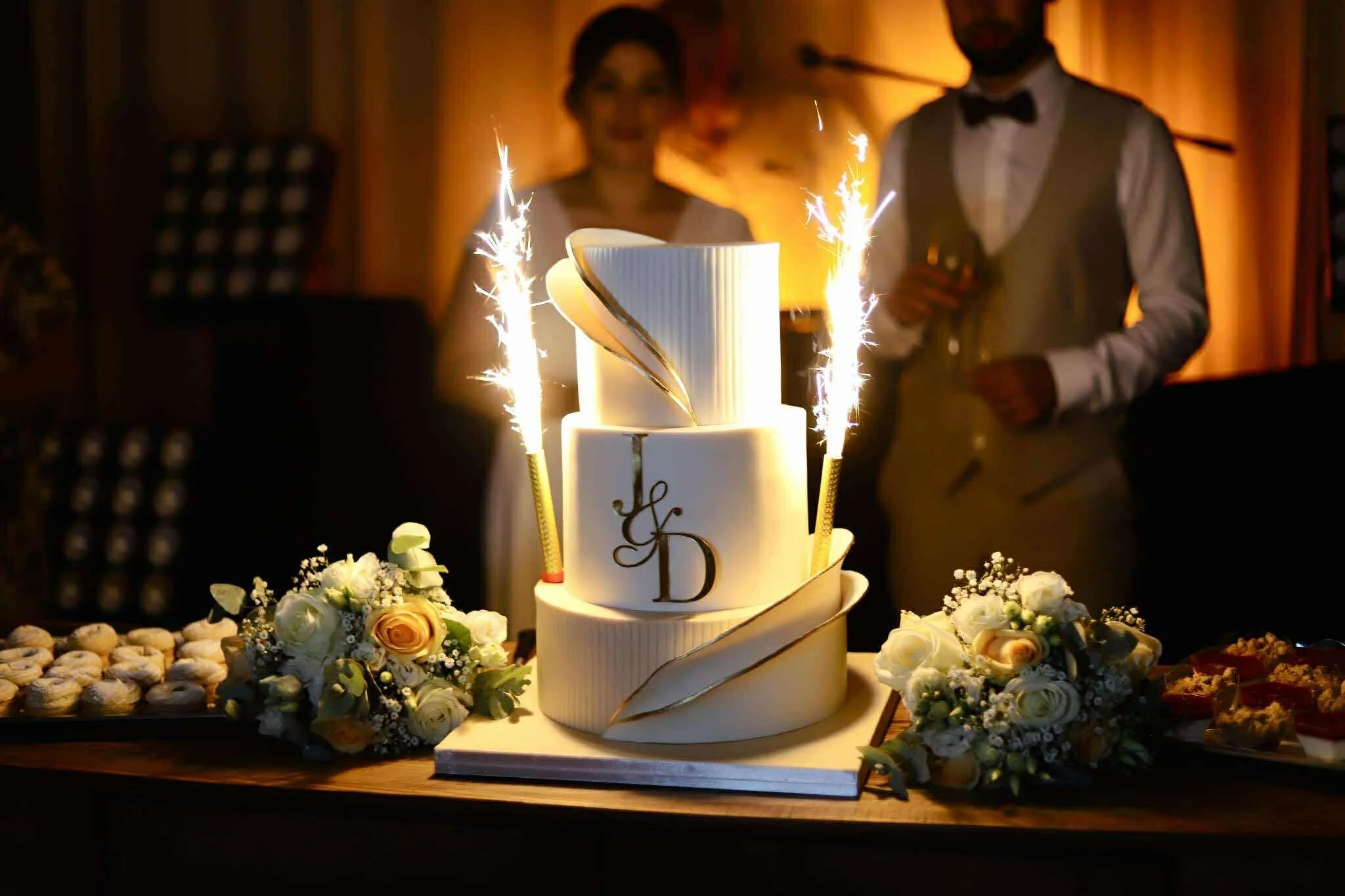 Gâteau de mariage blanc avec des décorations dorées, flammes d'étincelles sur le dessus, entouré de bouquets de fleurs blanches et jaunes, avec deux personnes en arrière-plan lors d'une célébration