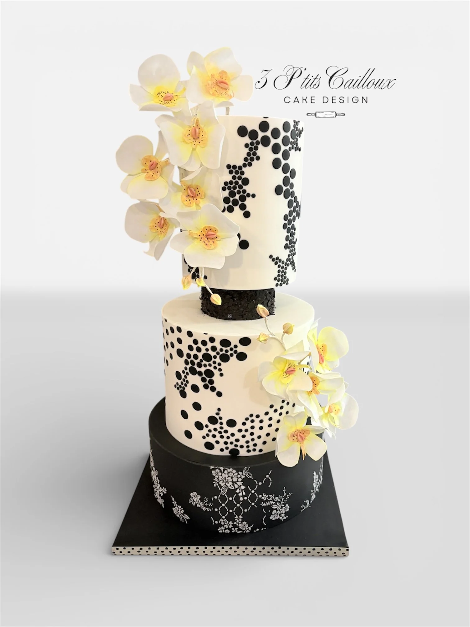 Gâteau à trois niveaux avec décoration florale d'orchidées blanches et jaunes, motifs noirs et blancs, et inscription '3 P'tits Cailloux'