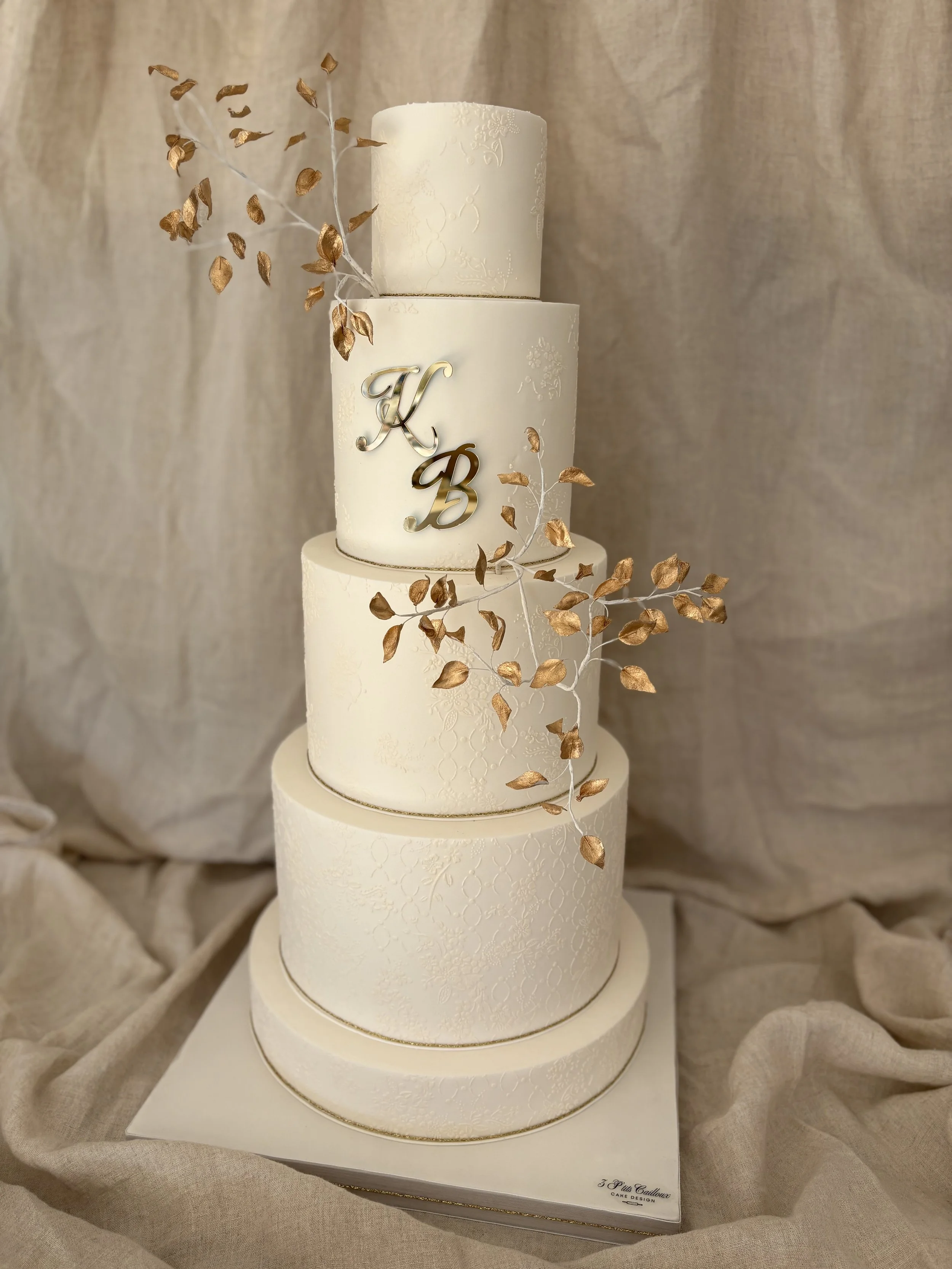 Gâteau de mariage à quatre niveaux blanc orné de branches de feuilles dorées et initiales en or K et B au centre.