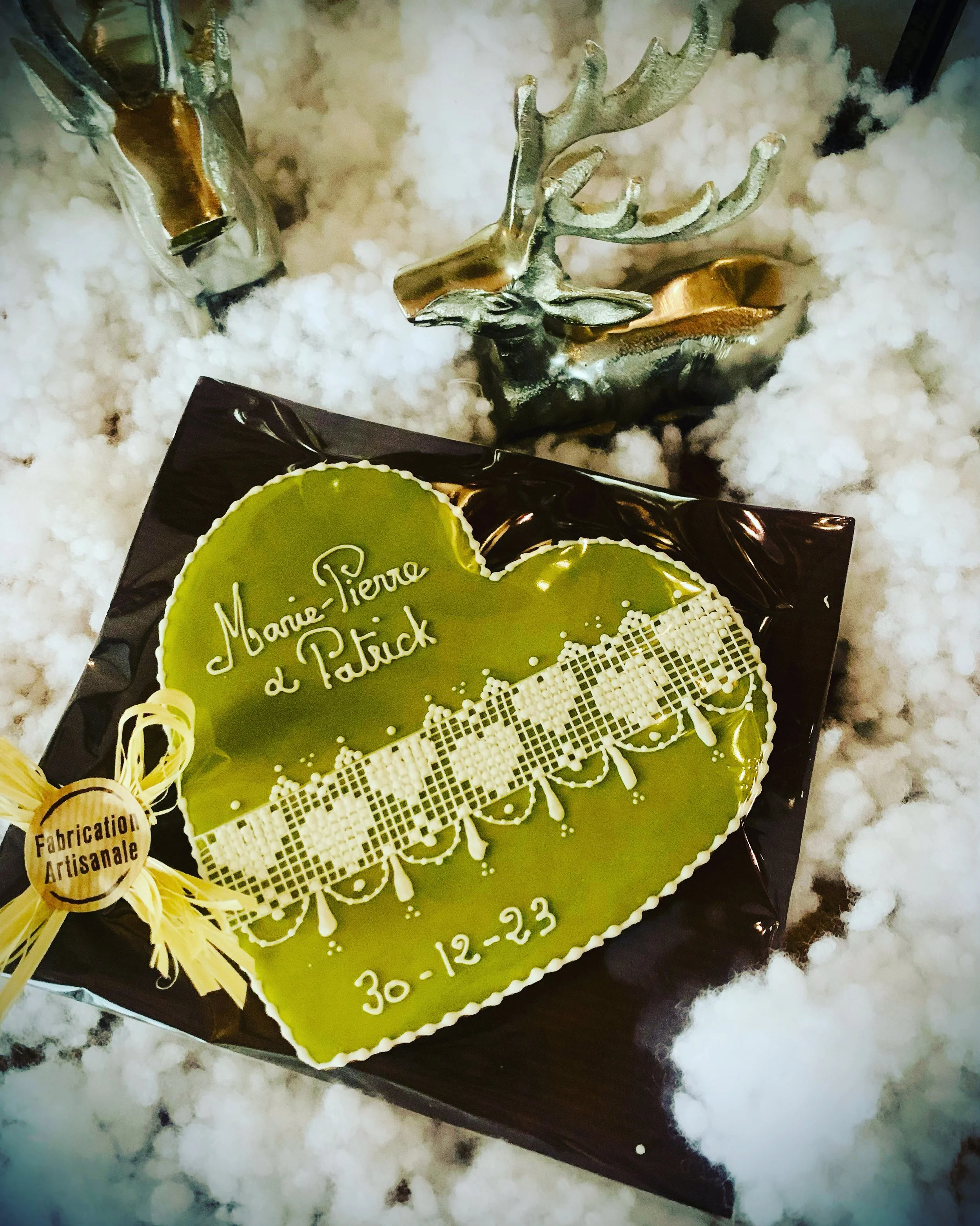 Un gâteau en forme de cœur vert avec une décoration en dentelle blanche et une inscription en français. Autour, des décorations de Noël, dont un renne doré et des éléments en or, sur un fond de coton ressemblant à de la neige.
