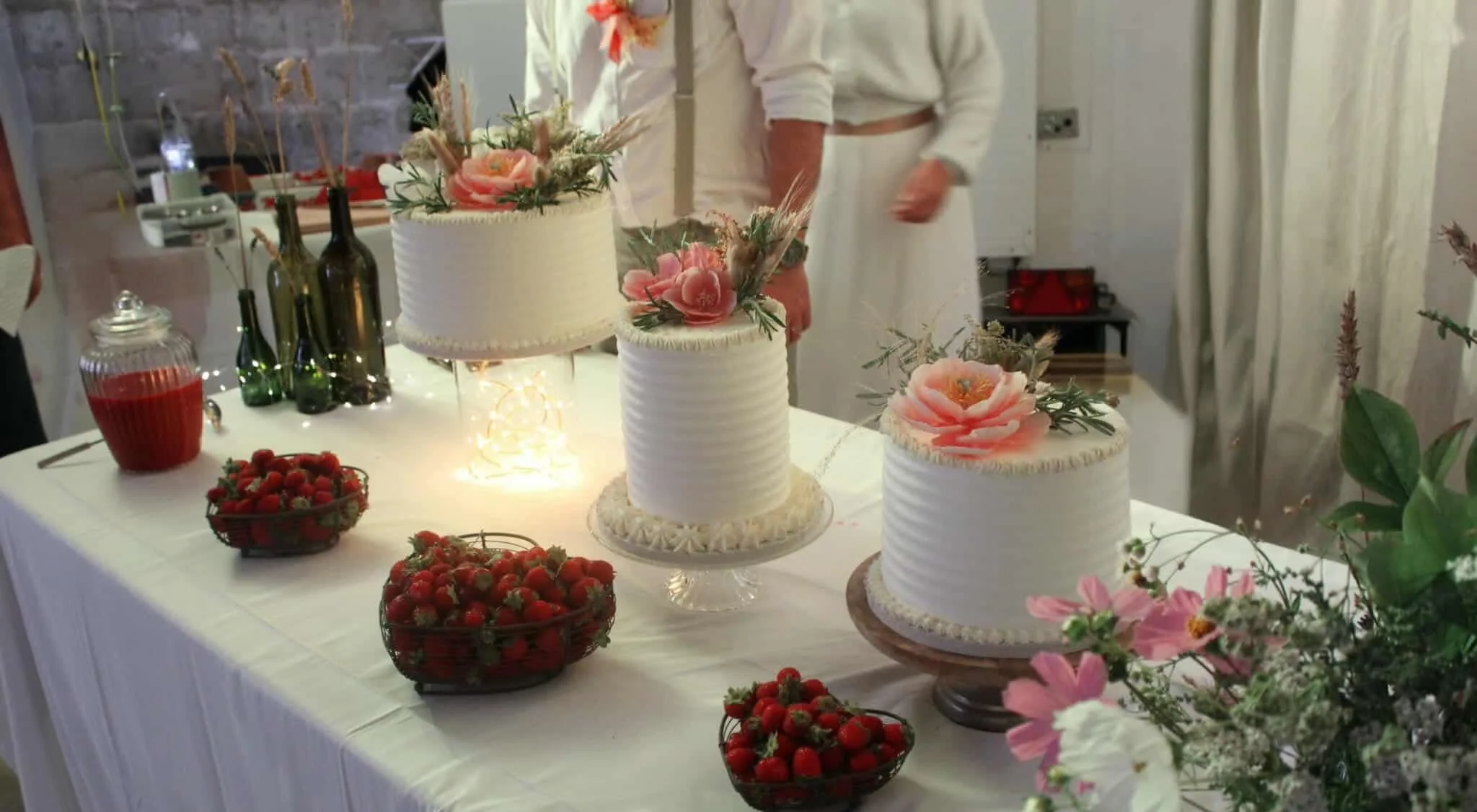 Table de fête avec trois gâteaux à la crème décorés de fleurs roses, plusieurs bols de fraises, et des bouteilles de vin ou d'huile, dans un cadre intérieur avec des personnes en arrière-plan.