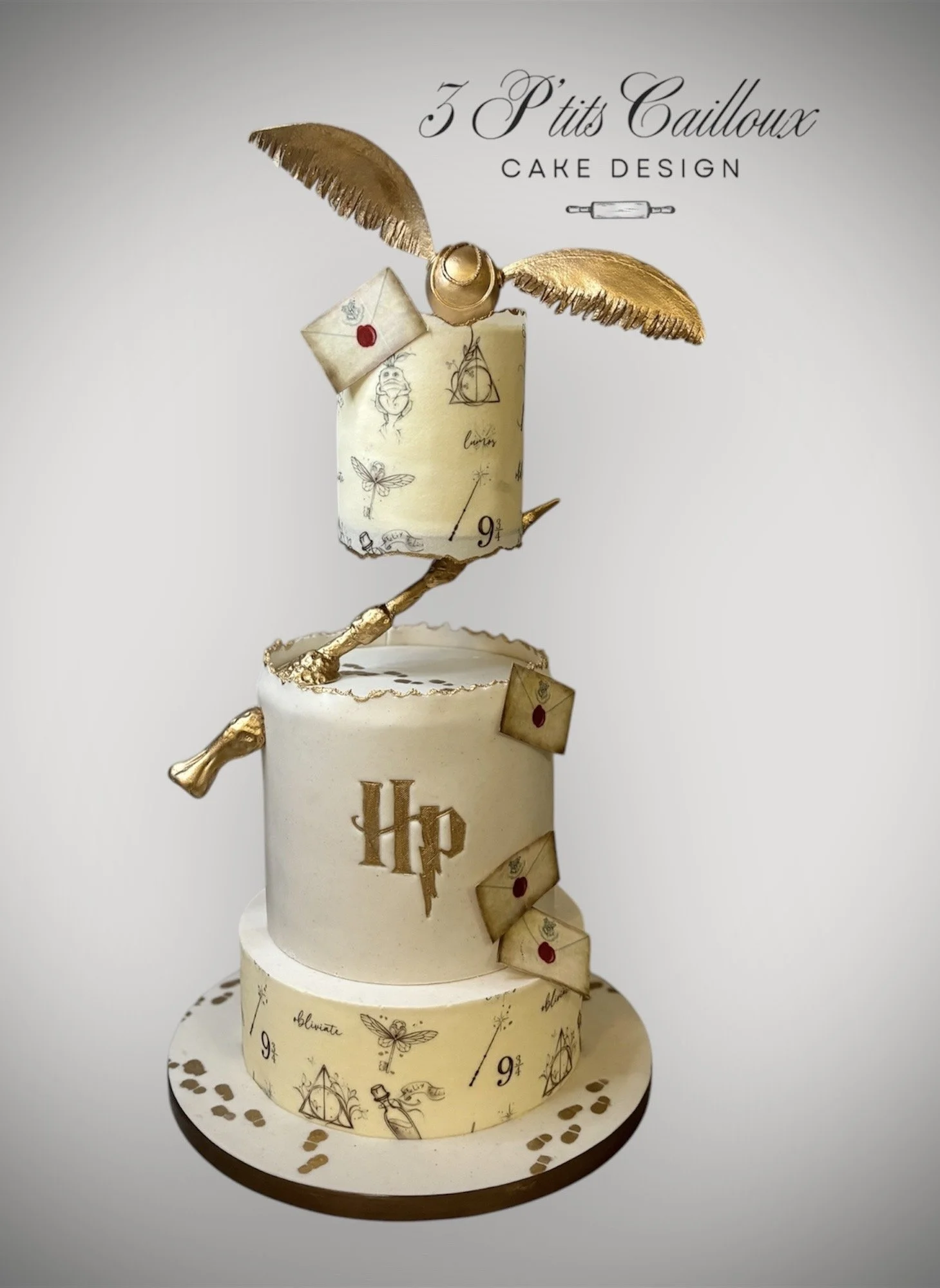 Gâteau décoré avec le thème Harry Potter, comprenant deux niveaux avec des éléments en or : une plume, une épée et des cartons portant des symboles de Harry Potter, avec le logo HP sur le deuxième niveau, et le texte '3 P'tits Cailloux' et 'Cake Desi