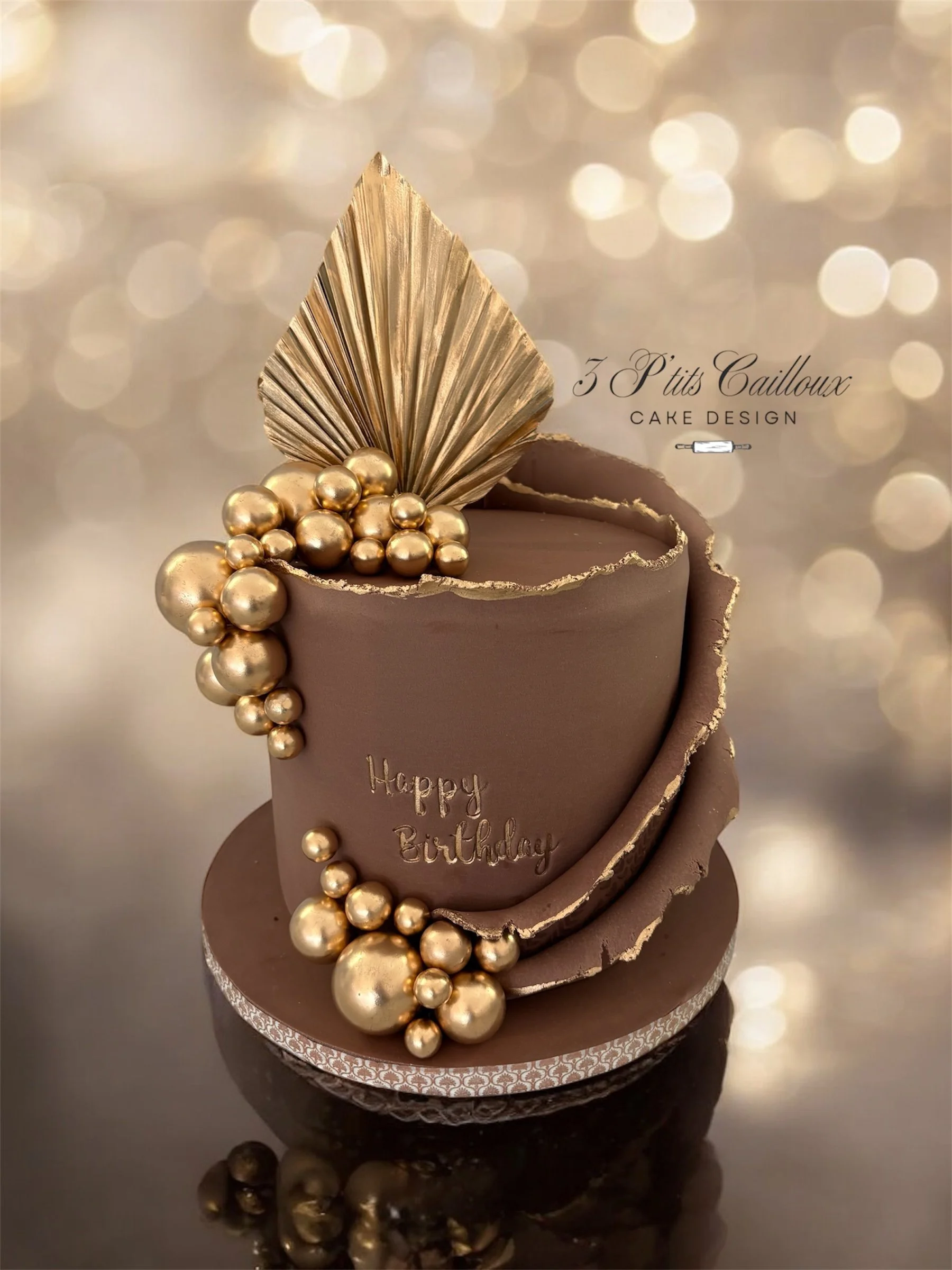 Un gâteau d'anniversaire au chocolat décoré de boules dorées et d'une grosse feuille dorée en style élégante, avec une inscription 'Happy Birthday' en gris. Le gâteau est placé sur une surface reflective, avec un fond flou lumineux.
