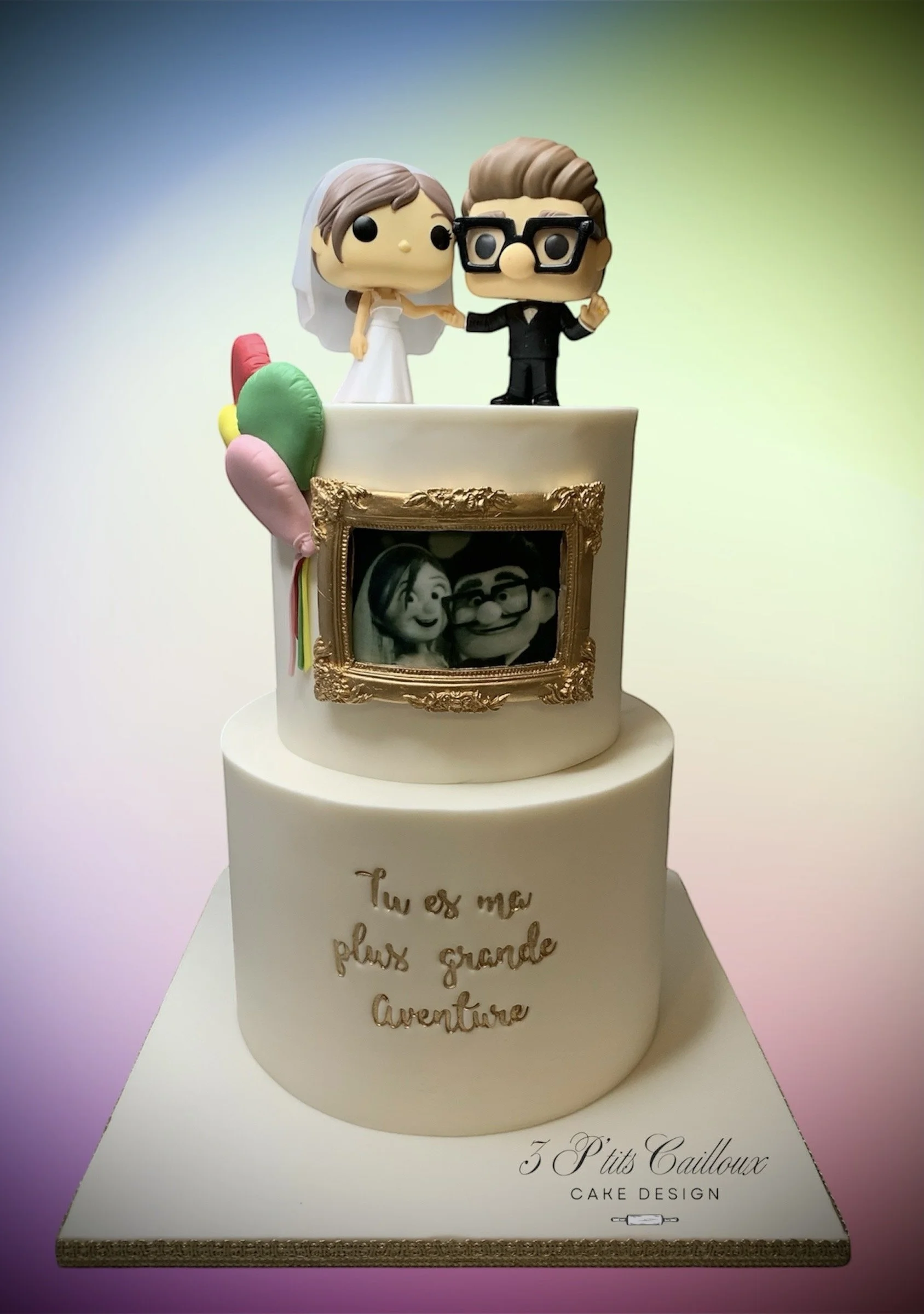 Gâteau de mariage avec des figurines de mariés et une photo en cadre, décorations colorées, inscription en français 'Tu es ma plus grande aventure', et une plaque mentionnant '3 P'tits Cailloux'.